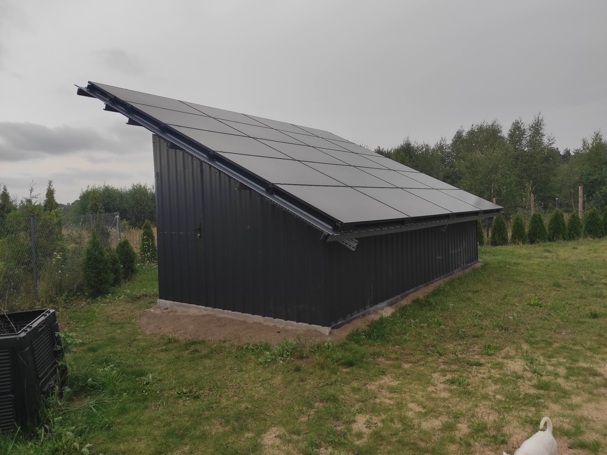 Czarny budynek gospodarczy z panelami fotowoltaicznymi na dachu, widoczny z zewnątrz na tle drzew i pochmurnego nieba. Z boku widoczny kompostownik i biały pies.