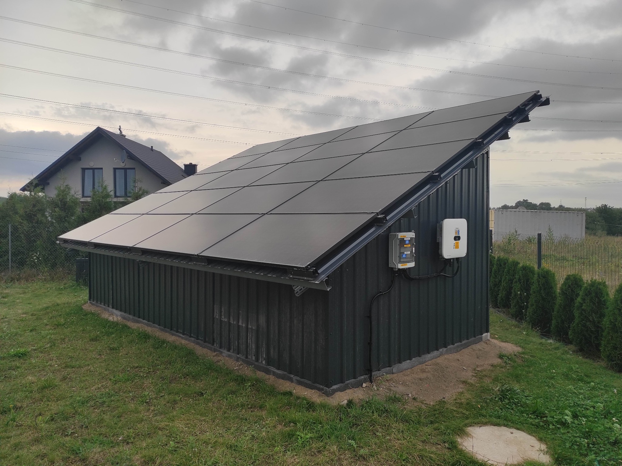 Mały budynek gospodarczy z panelami fotowoltaicznymi na dachu, inwerter zamontowany na ścianie, w tle dom jednorodzinny i pochmurne niebo.