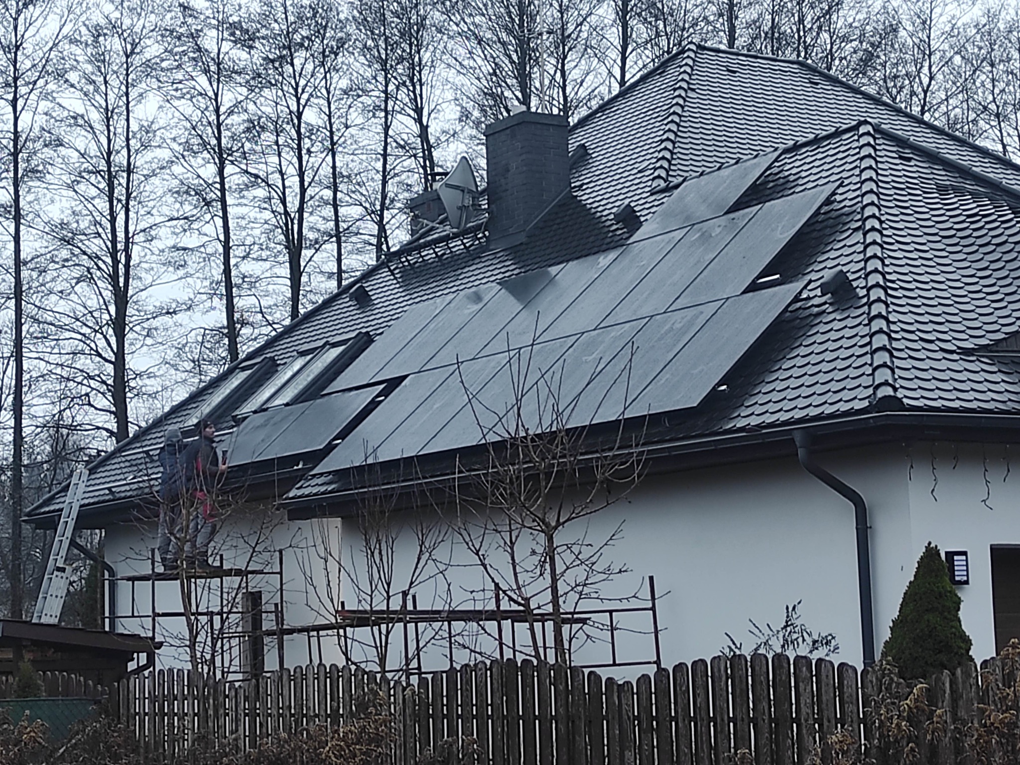 Montaż paneli słonecznych na dachu domu z dachówką łuskową; dwaj monterzy na rusztowaniu, drabina oparta o ścianę, drzewa w tle.