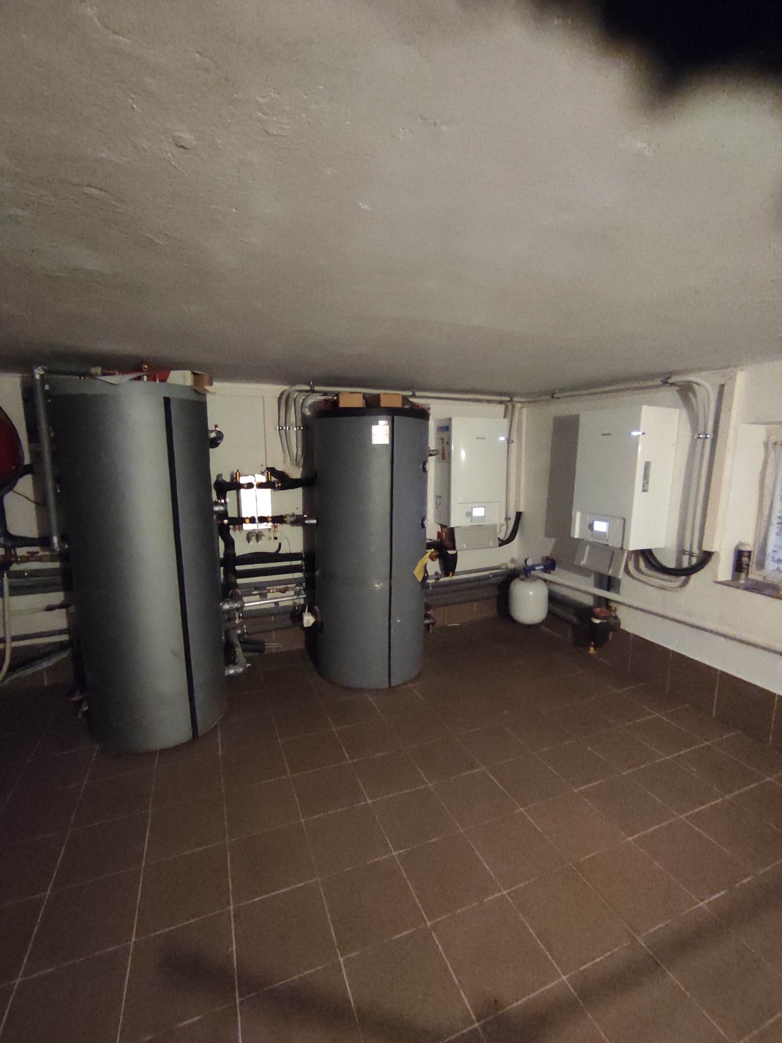 Dwa szare zbiorniki akumulacyjne ciepła w kotłowni z brązowymi płytkami podłogowymi, widoczne rury i zawory, oraz dwa białe piece wiszące na ścianie.