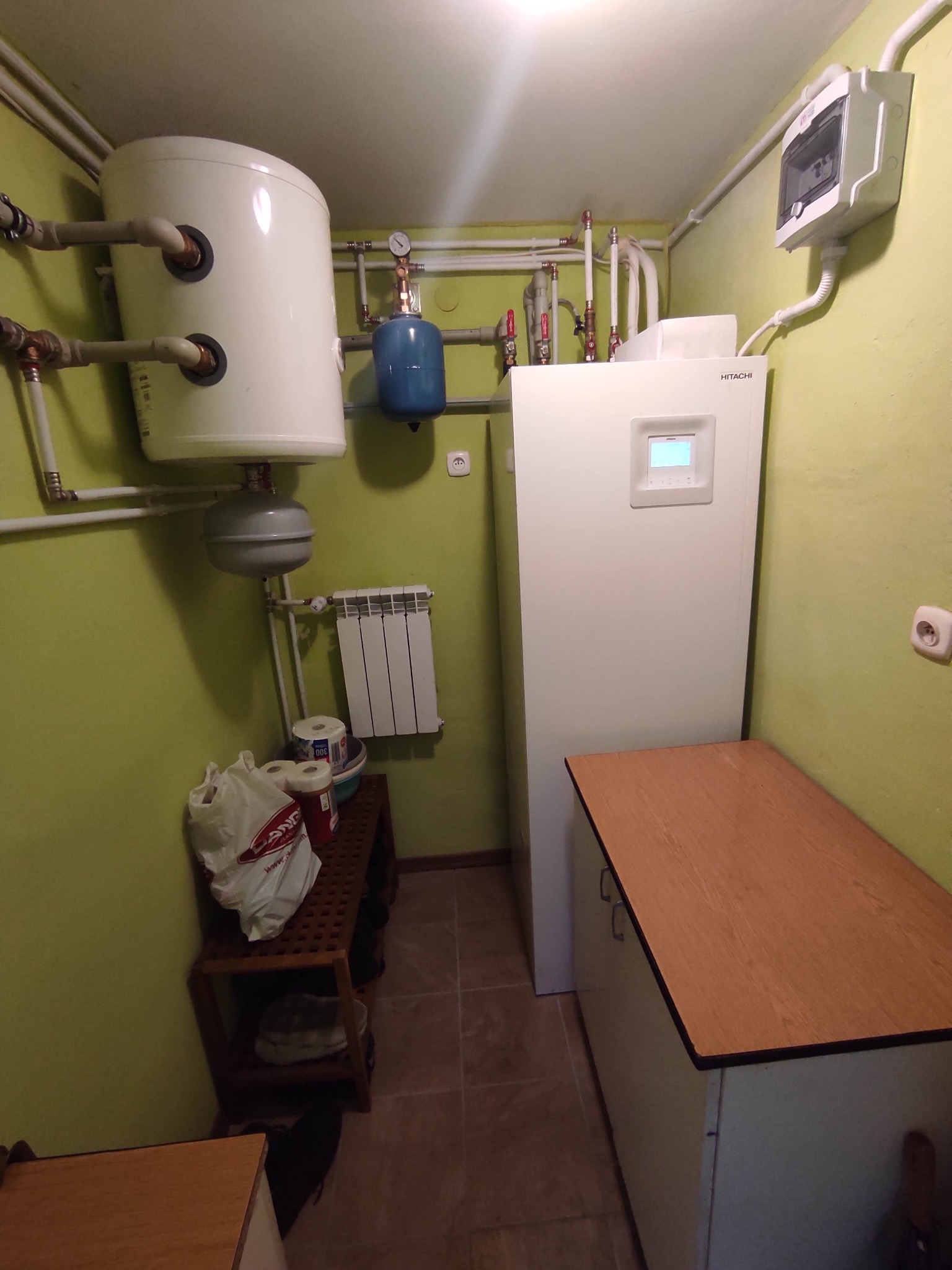 Kotłownia z białą pompą ciepła Hitachi, zbiornikiem wyrównawczym, grzejnikiem i szafką, widok na ścianę w odcieniu zieleni.