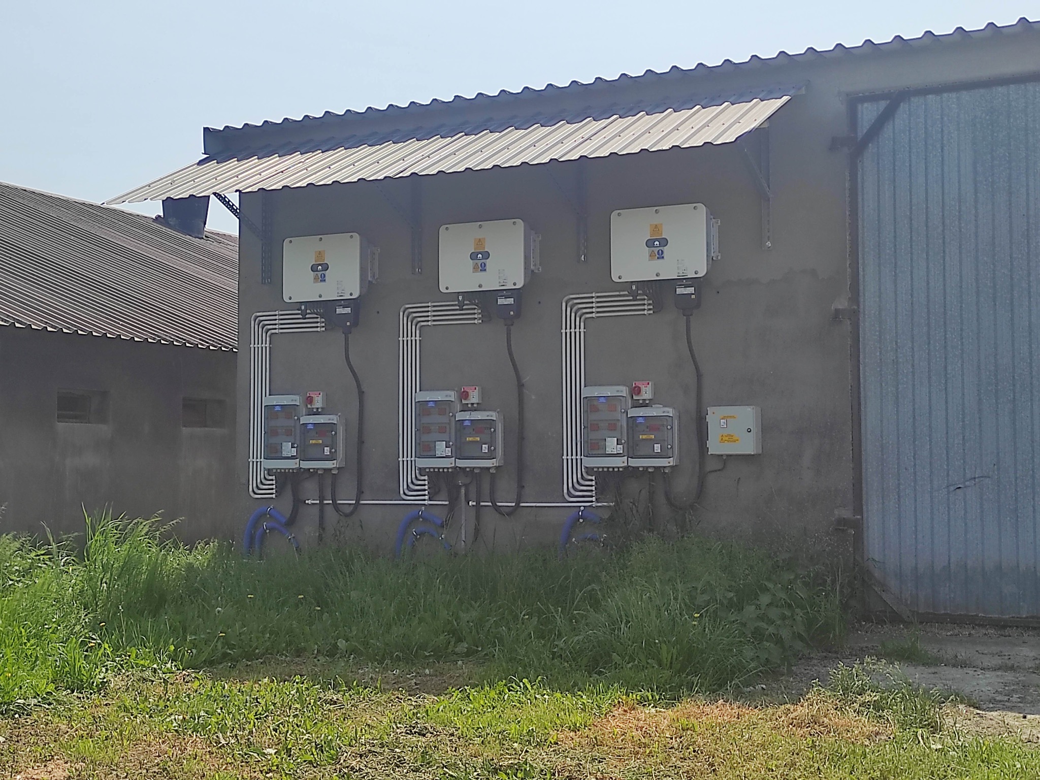 Trzy falowniki fotowoltaiczne zamontowane na szarej ścianie budynku gospodarczego z metalowym dachem, widoczne przewody w białych rurkach i niebieskie rury, zarośnięte trawą.