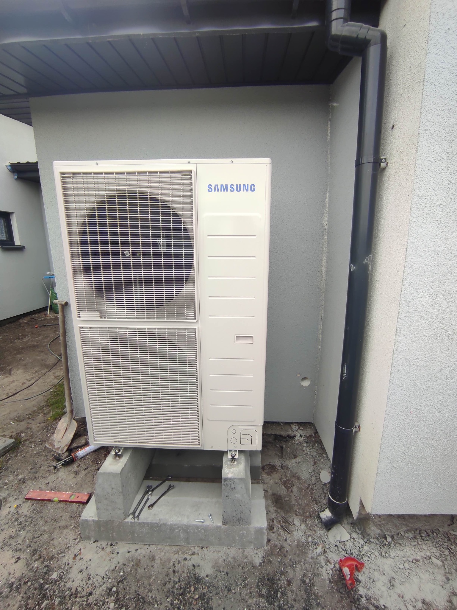 Biała pompa ciepła Samsung zamontowana na betonowej podstawie przy ścianie budynku z rurą spustową, widoczne narzędzia i poziomica sugerujące trwający montaż.