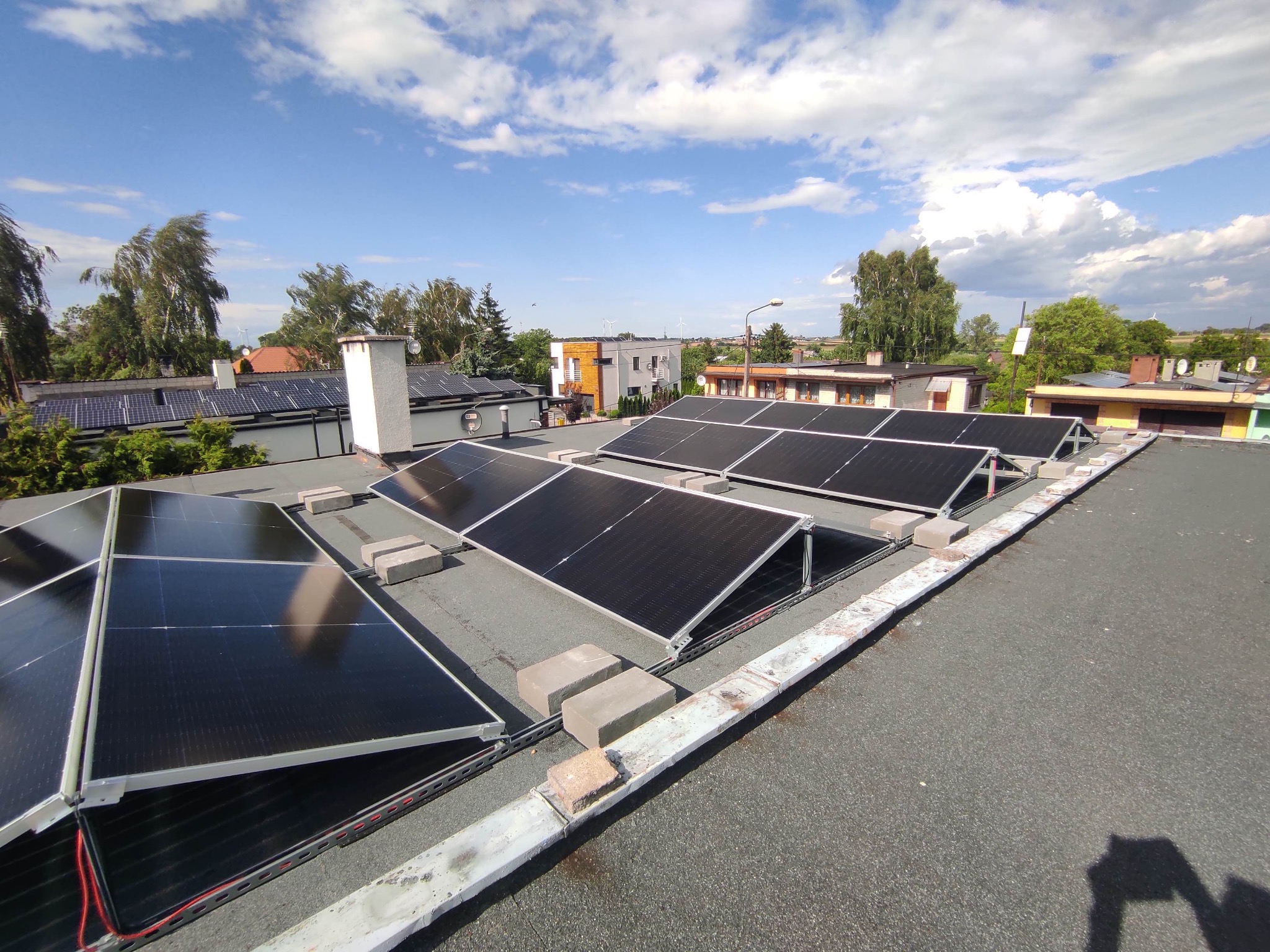 Instalacja fotowoltaiczna na płaskim dachu z widocznymi panelami, konstrukcją wsporczą i betonowymi bloczkami, w tle zabudowa jednorodzinna i błękitne niebo z chmurami.