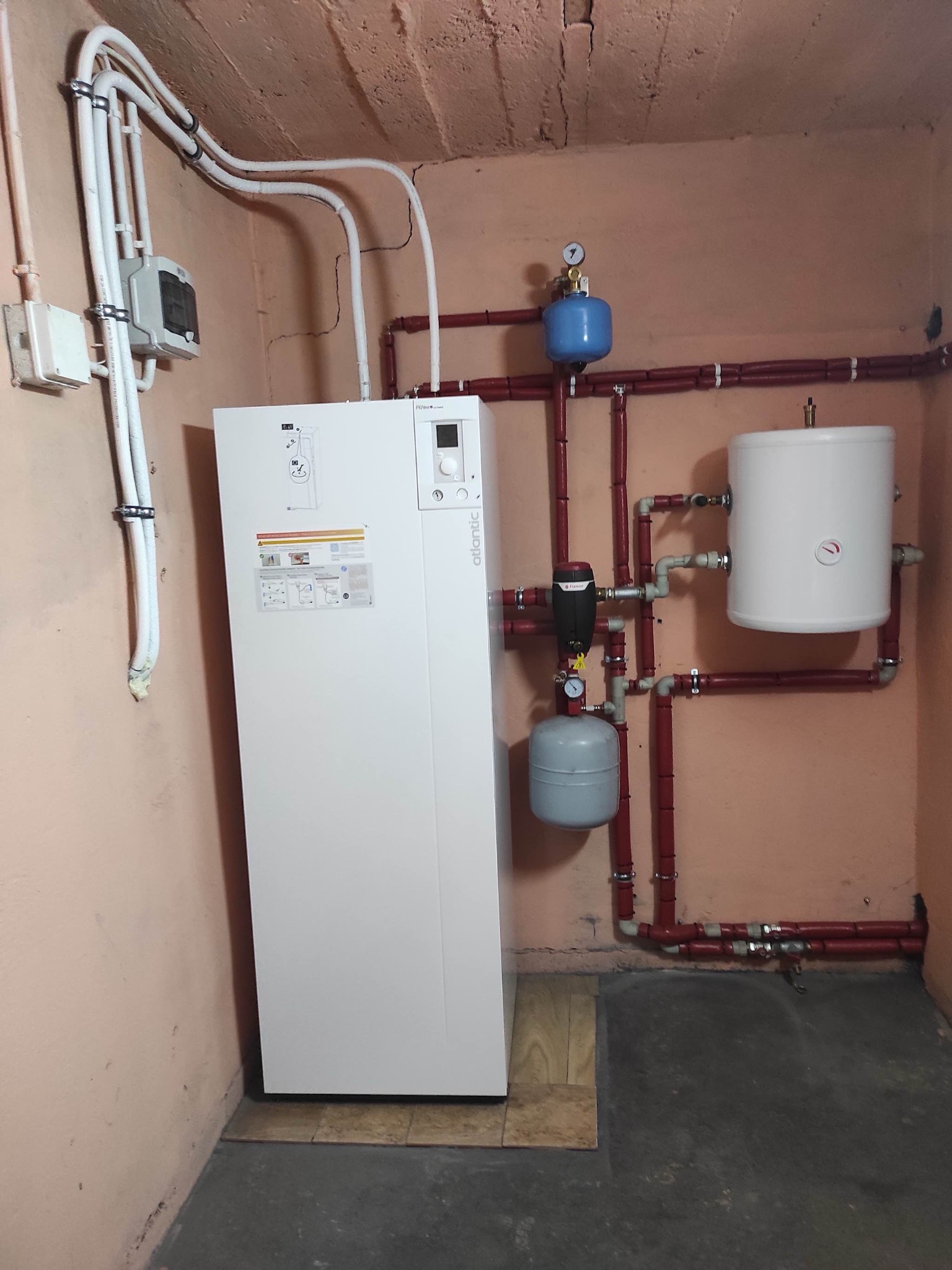 Biała pompa ciepła Atlantic w piwnicy z rurami w kolorze cegły, zbiornikiem wyrównawczym i manometrem.