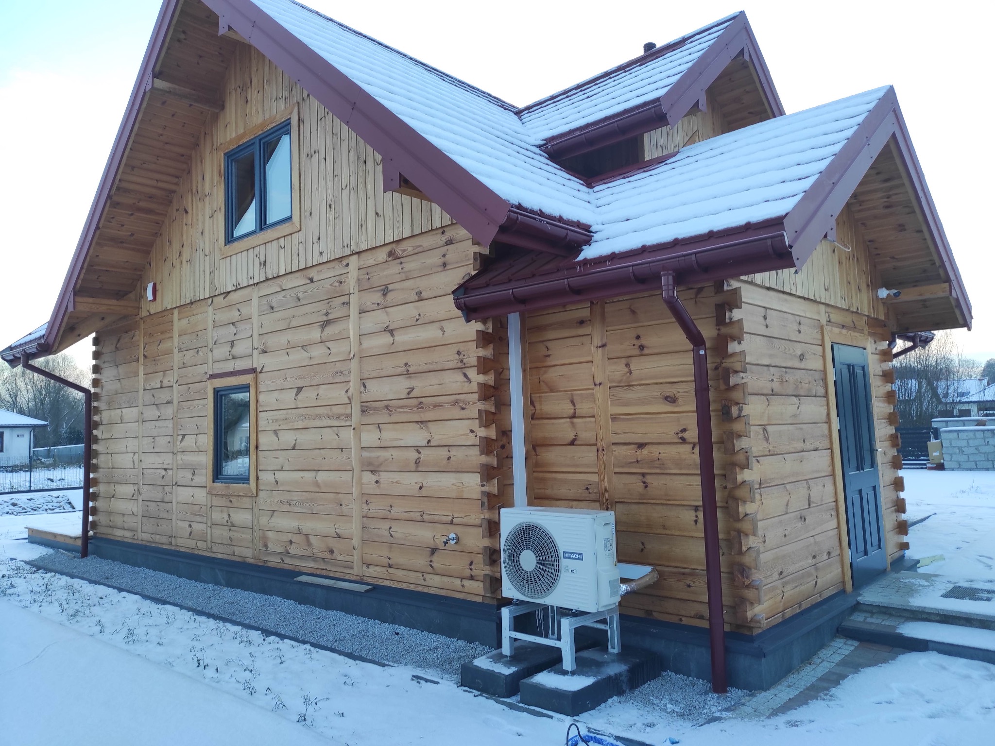 Zewnętrzna jednostka klimatyzacji Hitachi zamontowana na metalowym stelażu przy drewnianym domu z brązowym dachem pokrytym śniegiem, widoczne rynny i fragment fundamentu obsypanego drobnym kamieniem.