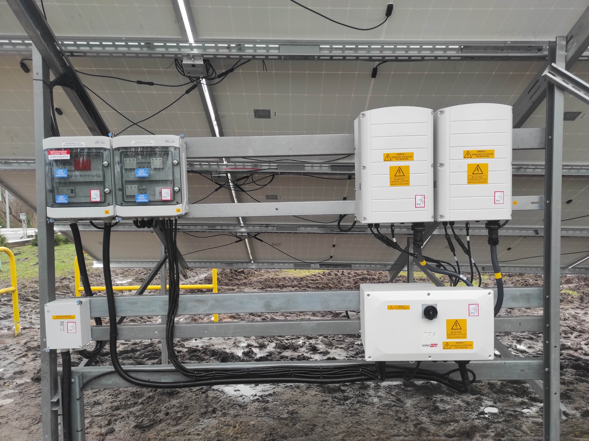 Widok od tyłu na instalację fotowoltaiczną z widocznymi panelami, skrzynkami elektrycznymi i inwerterami zamontowanymi na metalowej konstrukcji, teren wokół jest błotnisty, co sugeruje niedawno...