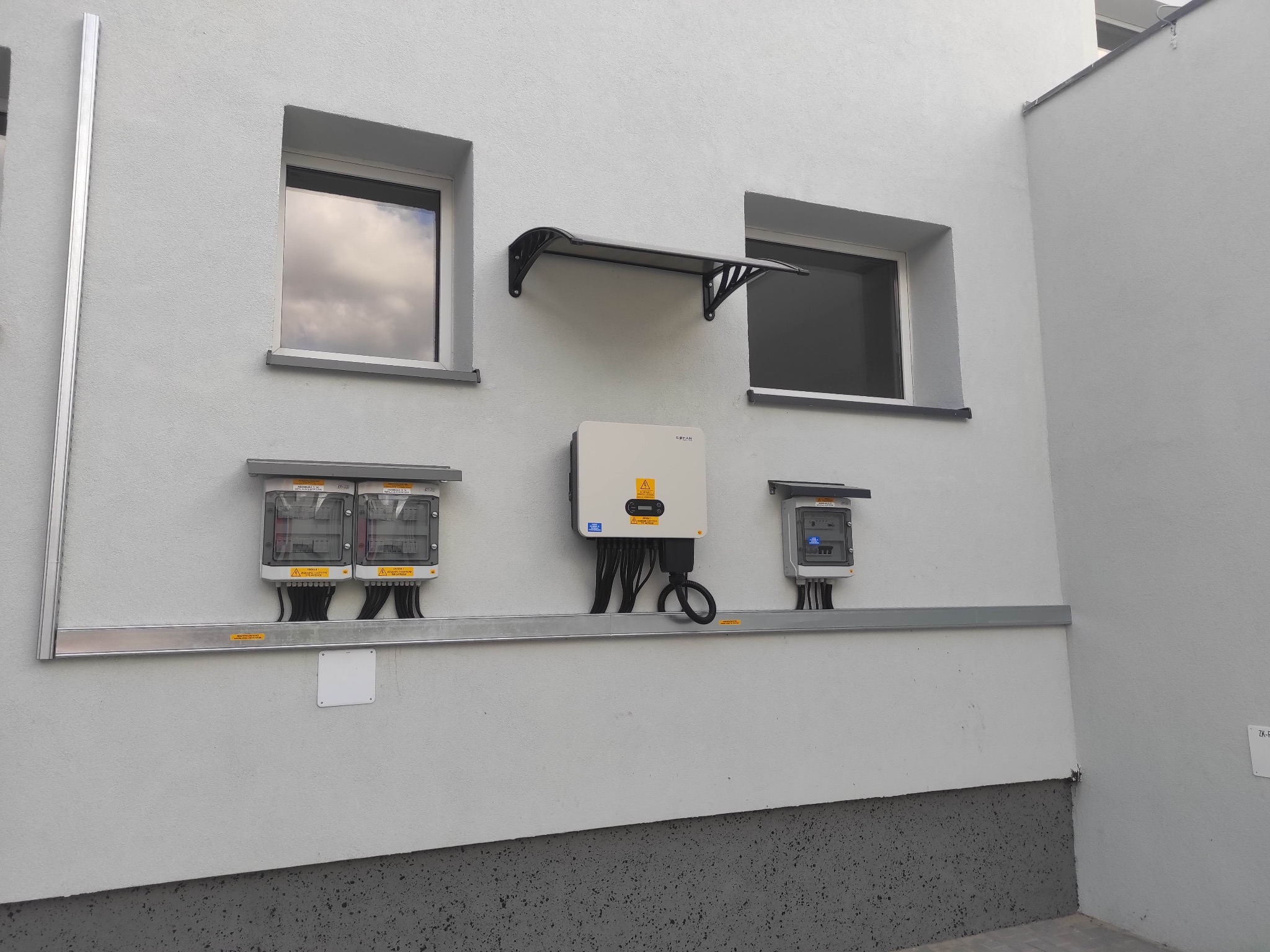 Biała ściana budynku z zamontowanymi elementami instalacji fotowoltaicznej: falownik, dwa liczniki, zabezpieczenia. Widoczne okna i daszek nad jednym z nich. Kabel z falownika zwinięty w pętlę.