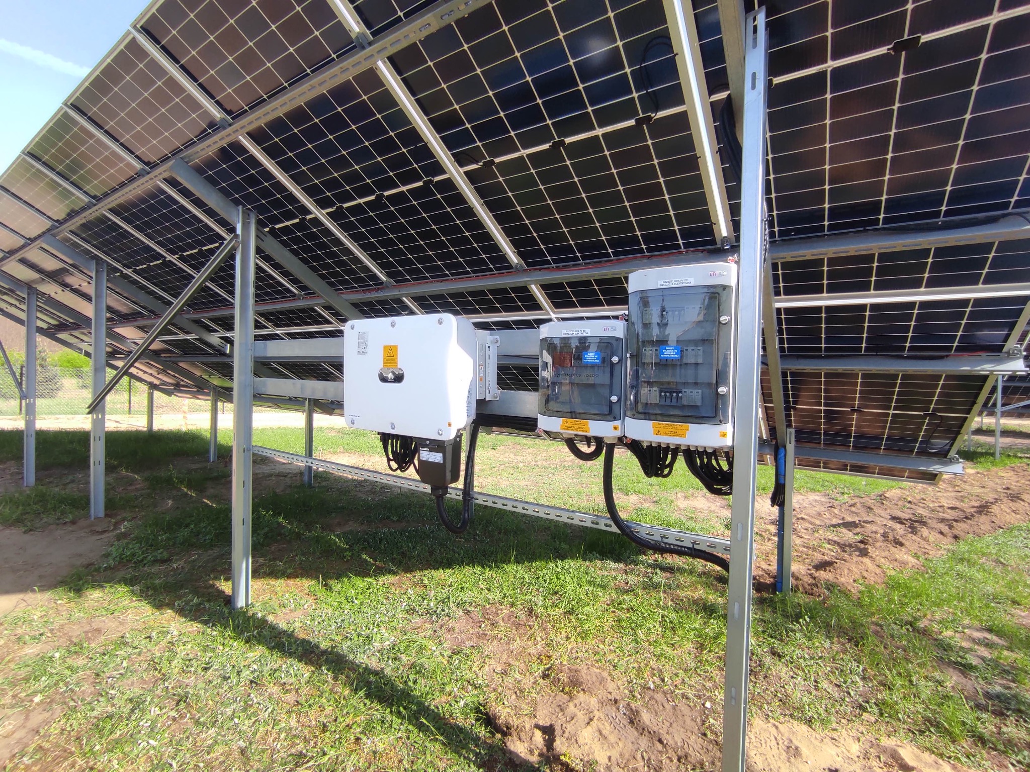 Widok od dołu na instalację fotowoltaiczną z widocznymi panelami, inwerterem i skrzynkami zabezpieczeń na tle trawy.