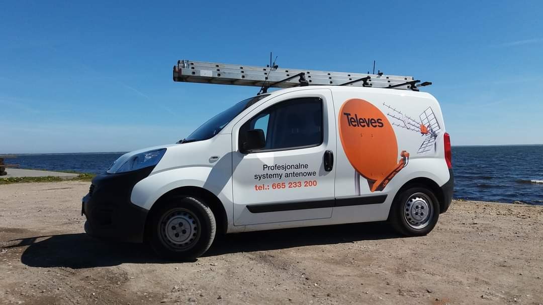 Biały samochód dostawczy z drabiną na dachu i logo Televes na tle błękitnego nieba i morza. Na boku pojazdu napis: Profesjonalne systemy antenowe tel.: 665 233 200.