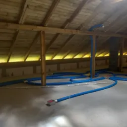 Niebieskie rury rekuperacji rozprowadzone na poddaszu z drewnianą konstrukcją dachu i ścianami z betonowych bloczków.