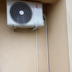 Zewnętrzna jednostka klimatyzatora Rotenso IMOTO zamontowana na beżowej elewacji budynku z widocznymi białymi rurami instalacyjnymi.