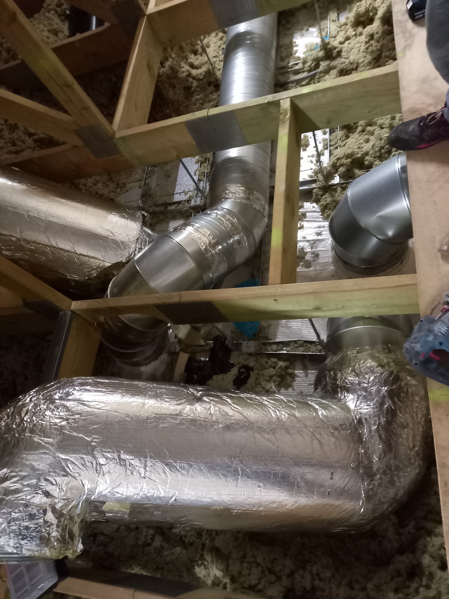 Instalacja systemu wentylacji mechanicznej z rekuperacją w przestrzeni poddasza, widoczne rury izolowane folią aluminiową, elementy drewnianej konstrukcji dachu oraz warstwa wełny mineralnej.