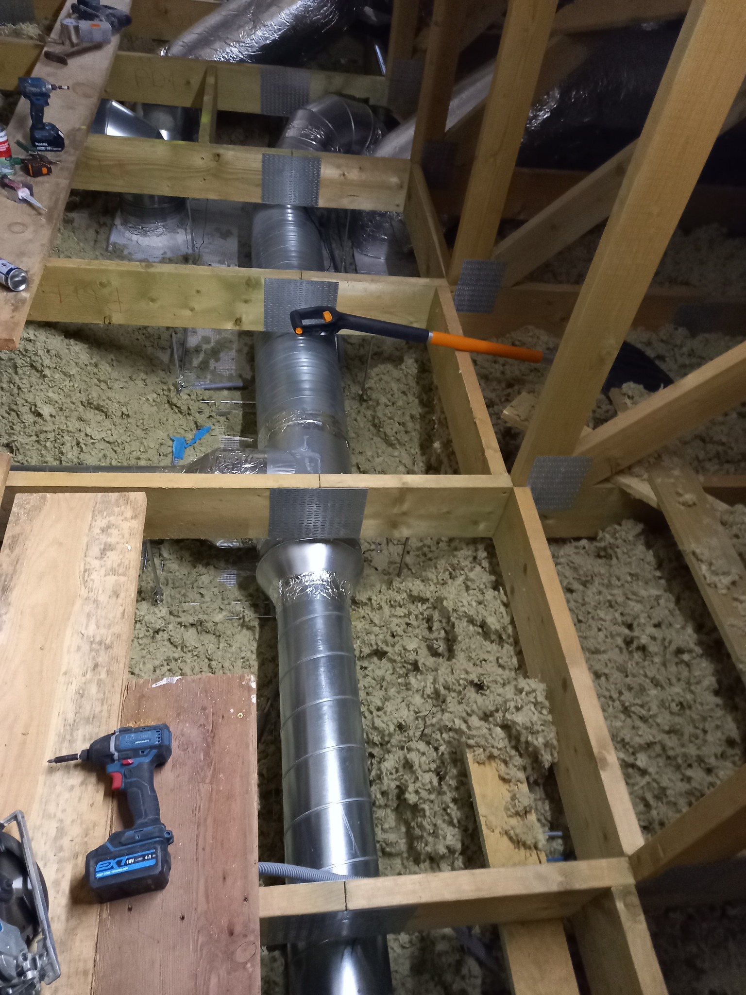 Montaż srebrnej, spiralnej rury wentylacyjnej w drewnianej konstrukcji poddasza, widoczna izolacja z wełny mineralnej, narzędzia pracy: wkrętarka i piła tarczowa.