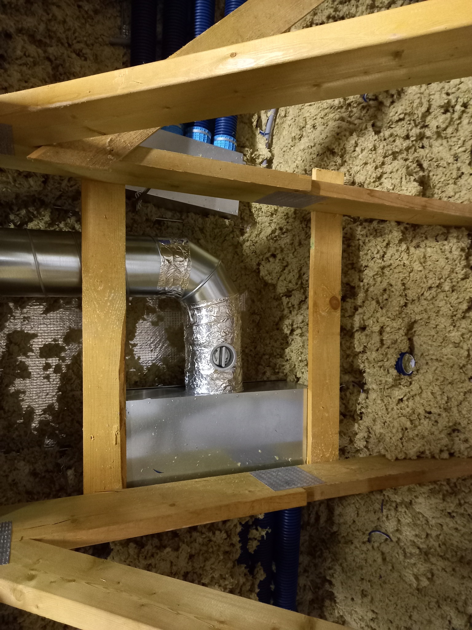 Fragment instalacji rekuperacji w przestrzeni poddasza z widocznymi elementami izolacji, drewnianej konstrukcji nośnej oraz przewodami wentylacyjnymi, zabezpieczonymi taśmą aluminiową.