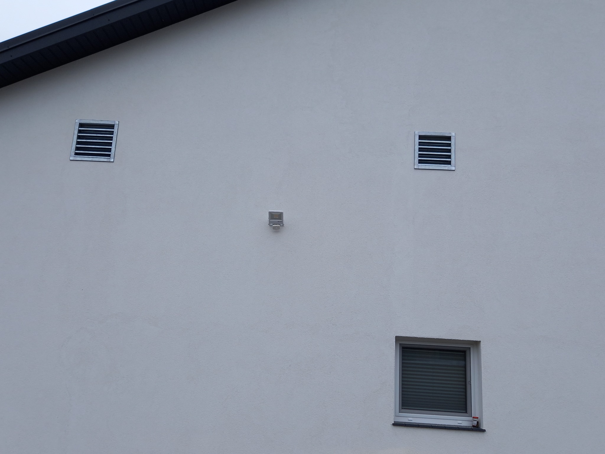 Elewacja budynku z dwoma kratkami wentylacyjnymi, lampą zewnętrzną i oknem z żaluzjami.