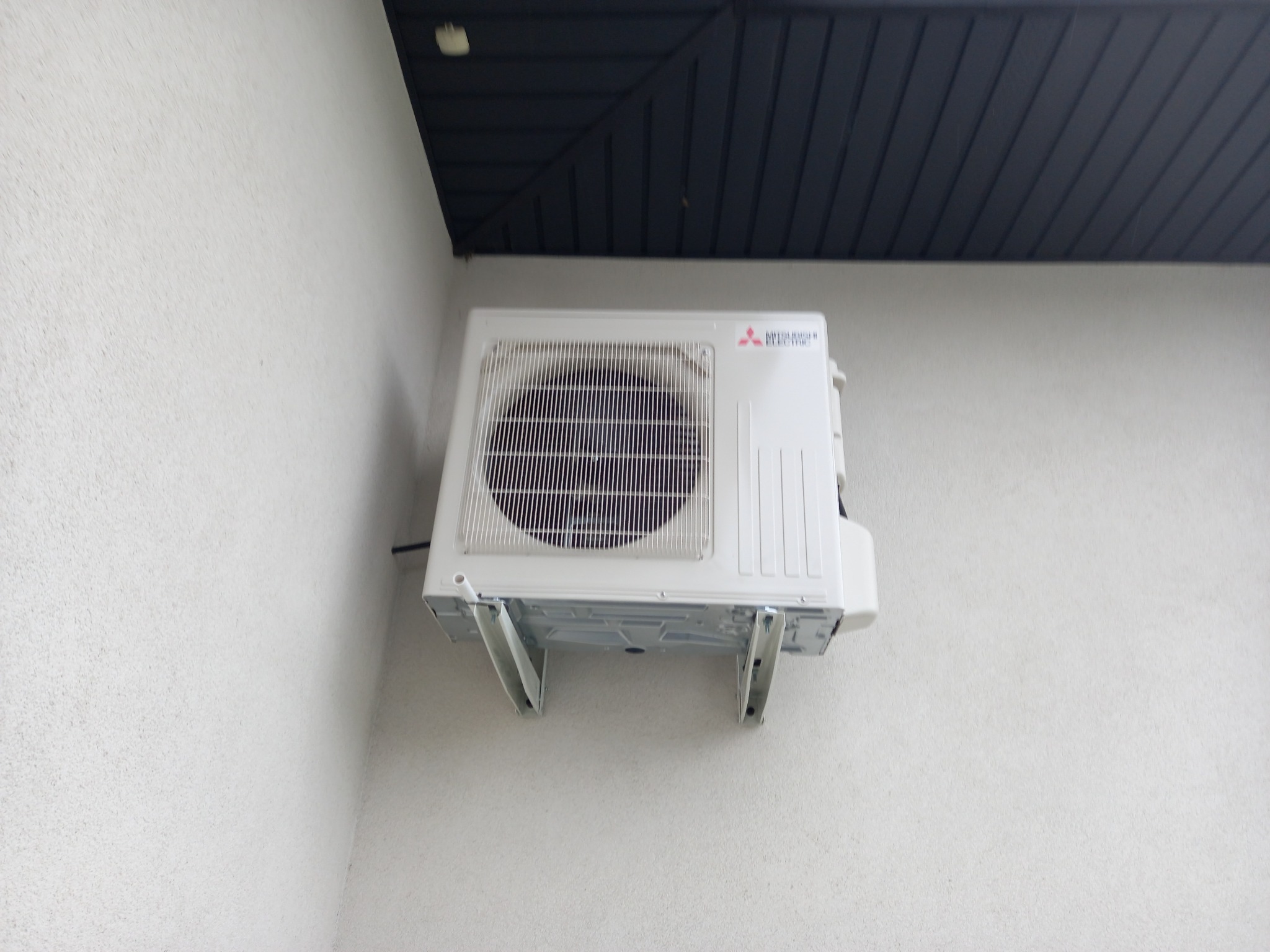 Biała jednostka zewnętrzna klimatyzatora Mitsubishi Electric zamontowana na ścianie budynku z szorstkim tynkiem, widoczna z dołu. Czarna podbitka dachowa w tle.