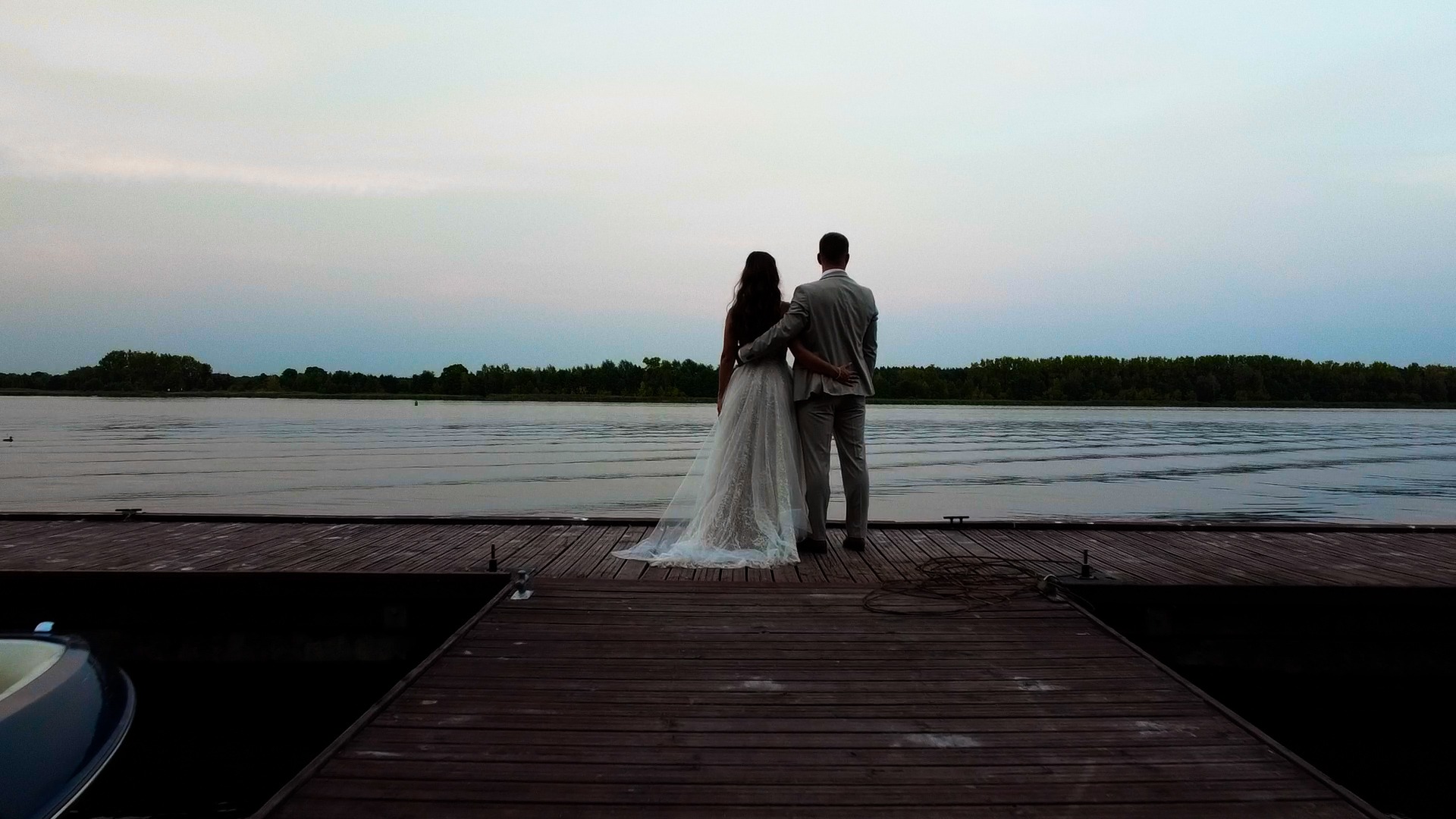 Para młoda, tyłem, na drewnianym molo nad jeziorem, w tle las i zachmurzone niebo o zmierzchu.