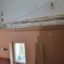 Daniel Janbrw - Wnętrze w trakcie remontu: ściany częściowo pokryte świeżym tynkiem, widoczne stare instalacje elektryczne, rury grzewcze i fragment okna z zielenią na zewnątrz.