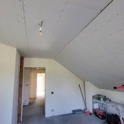 Daniel Janbrw - Poddasze w trakcie prac wykończeniowych: zamontowane płyty gipsowo-kartonowe na skosach i suficie, widoczny podnośnik do płyt, narzędzia na regale i wystające kable ze ściany.