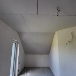 Pokój na poddaszu w trakcie przygotowywania do szpachlowania, z widocznymi płytami gipsowo-kartonowymi na suficie i ścianach, surowym betonem na podłodze, wiszącą żarówką na kablu oraz wystającymi...