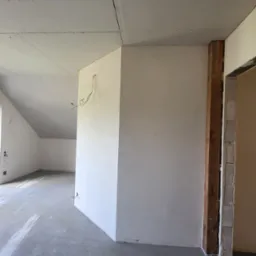 Surowe wnętrze poddasza z wykończonymi płytami gipsowo-kartonowymi na skosach i ścianach, widoczne kable elektryczne wystające ze ściany, surowa podłoga i fragment ościeżnicy drzwiowej.