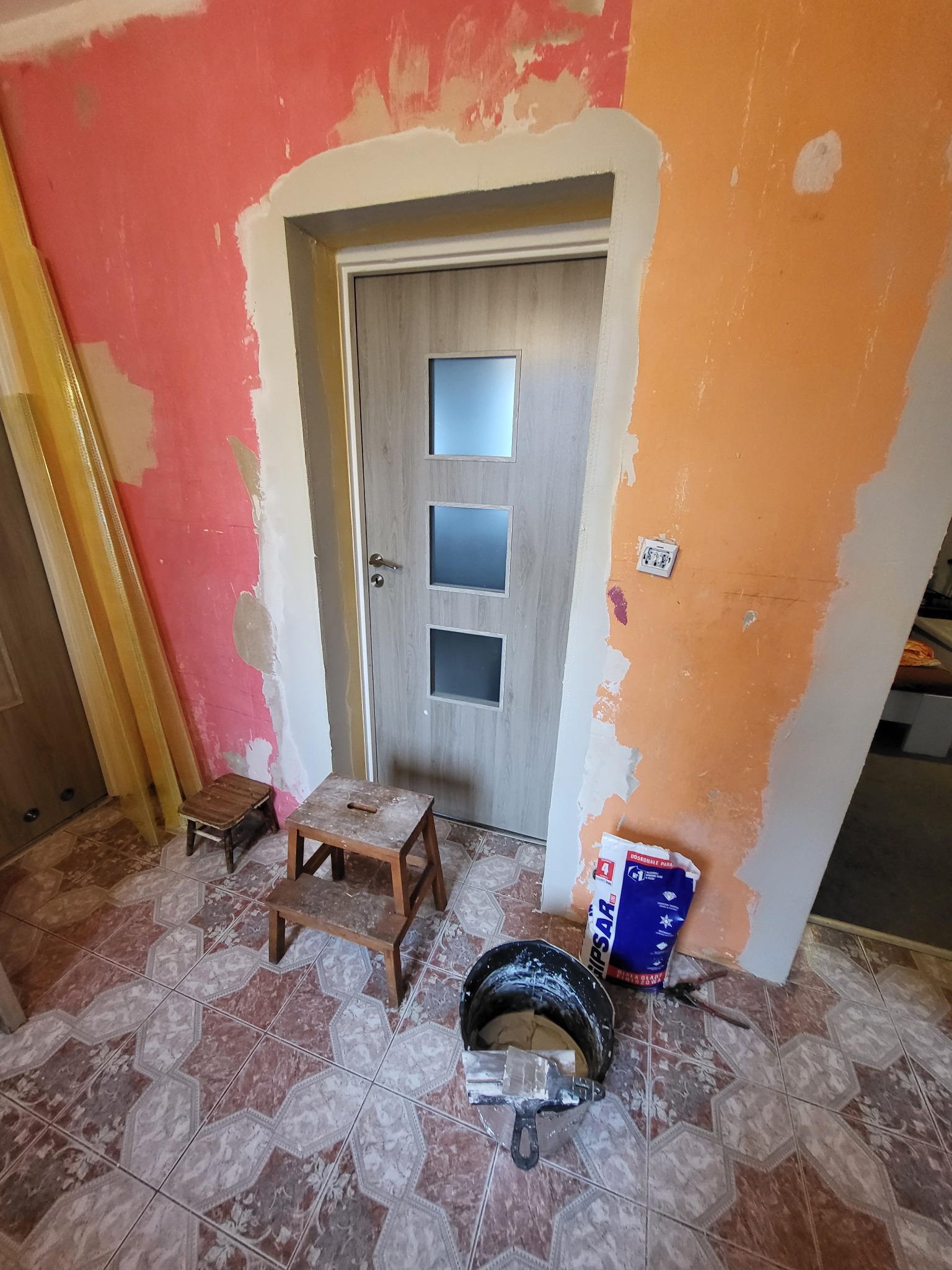Wnętrze w trakcie prac przygotowawczych do wykończenia, widoczne fragmenty ścian z nałożoną gładzią, zamontowane drzwi z trzema szybkami, wiadro z narzędziami i workiem gipsu na wzorzystej podłodze.