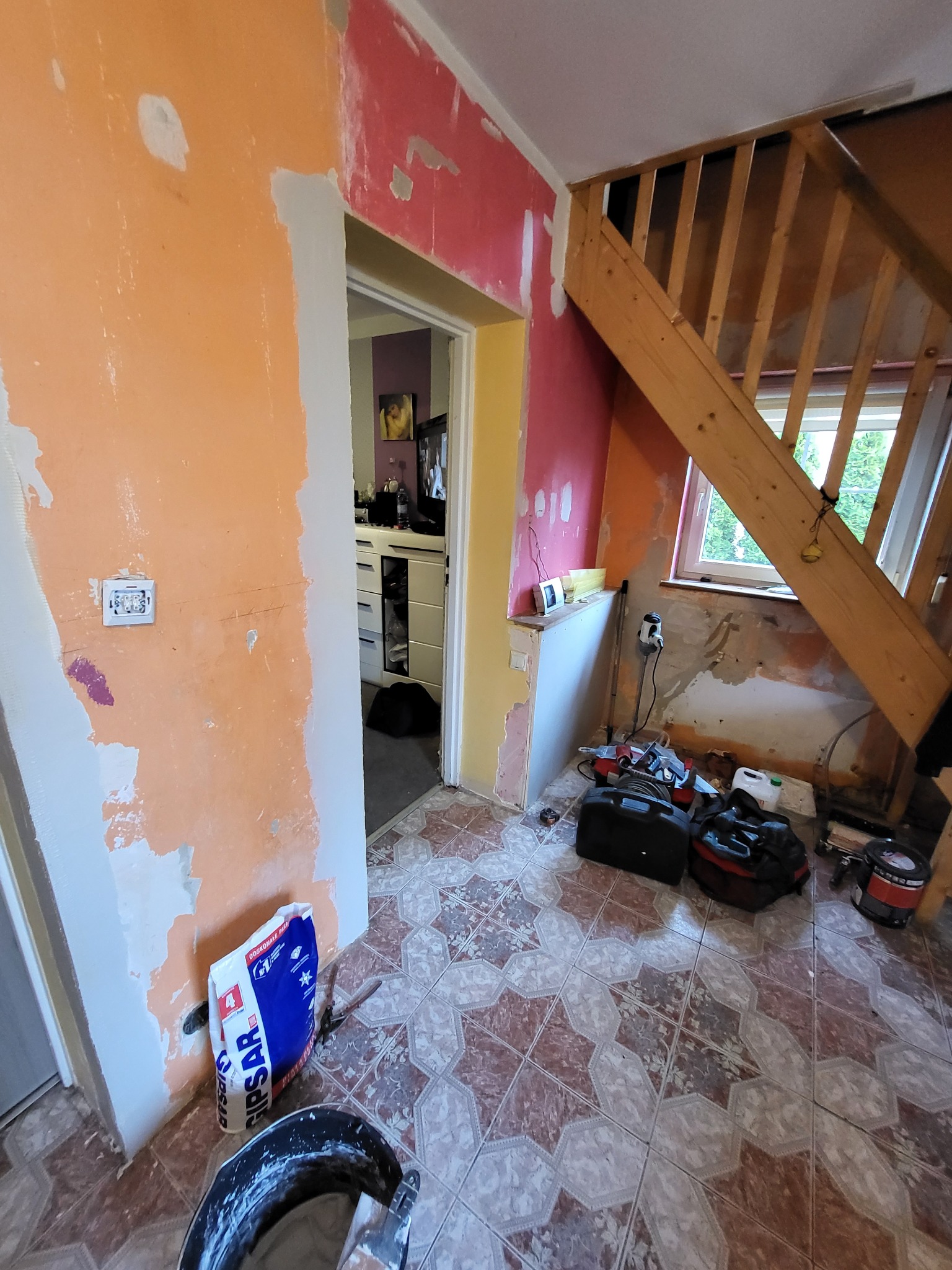 Wnętrze w trakcie przygotowania do malowania: ściany w kolorach pomarańczowym i różowym z widocznymi śladami po szpachlowaniu, drewniane schody, podłoga wyłożona płytkami w geometryczny wzór, torby...