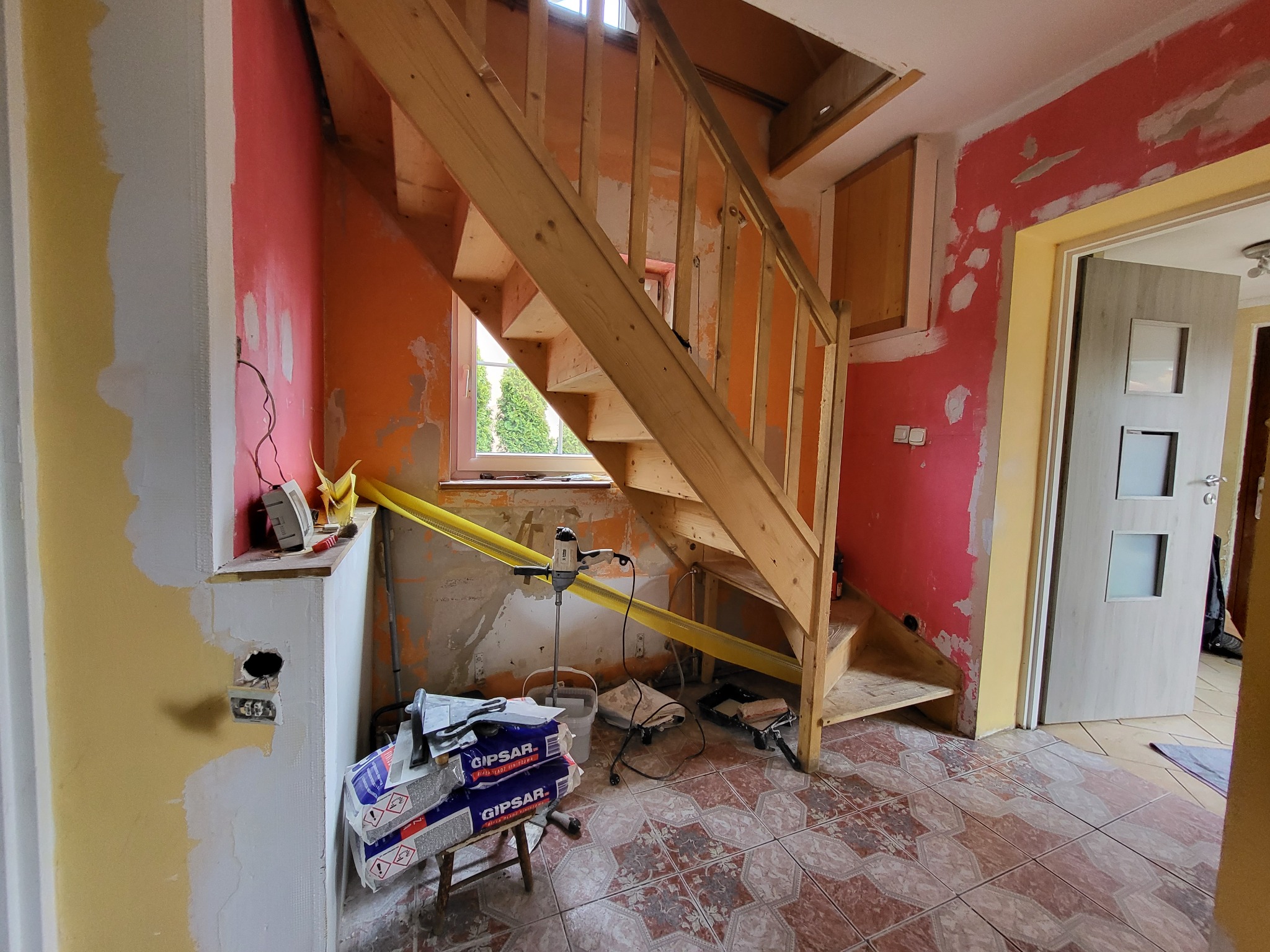Wnętrze domu w trakcie remontu: drewniane schody, ściany w trakcie szpachlowania, widoczne worki z gipsem i narzędzia.