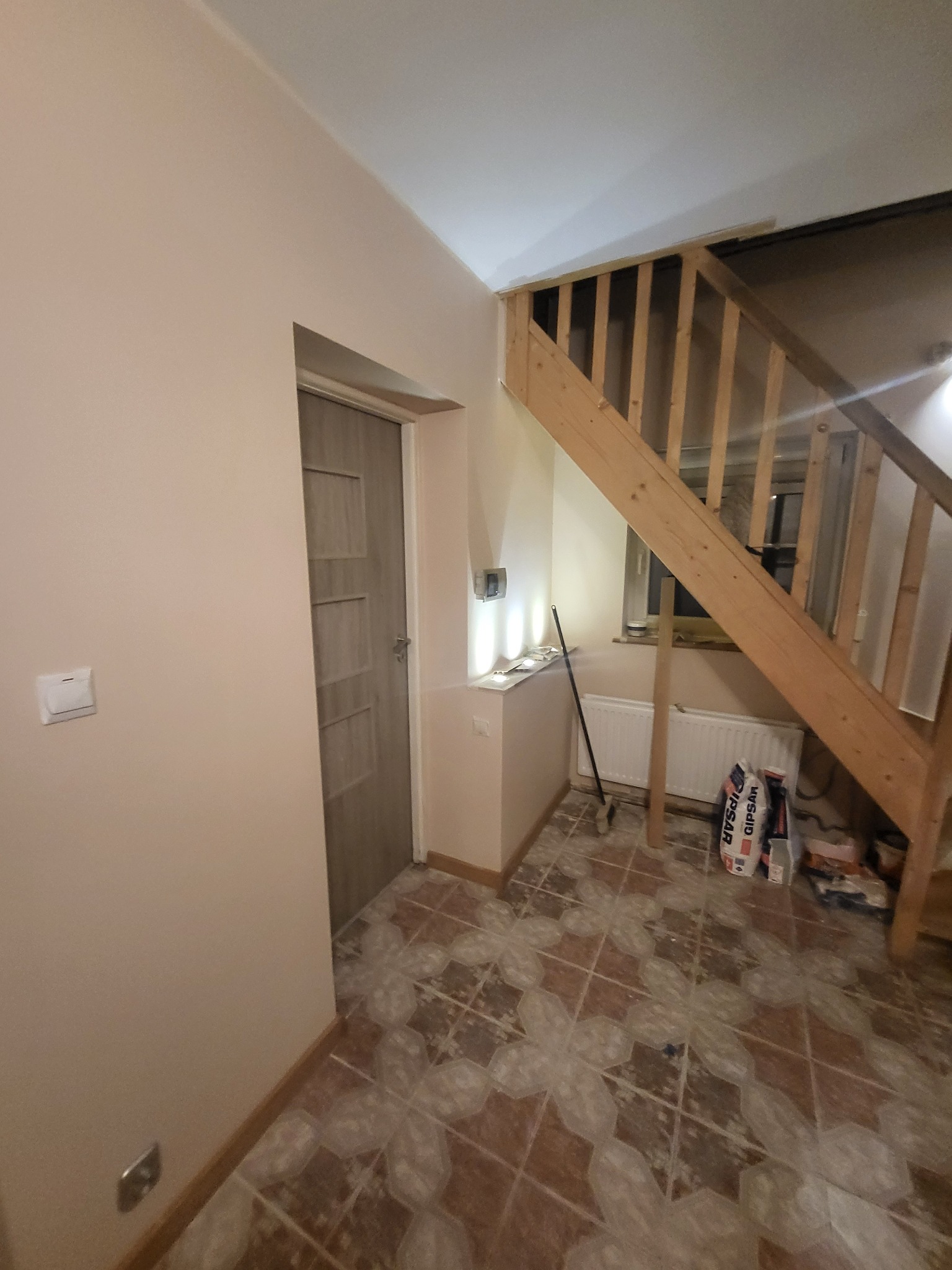 Wnętrze domu w trakcie remontu: widoczne schody drewniane, drzwi wewnętrzne, ściany po szpachlowaniu, podłoga zabezpieczona folią oraz worki z gipsem.