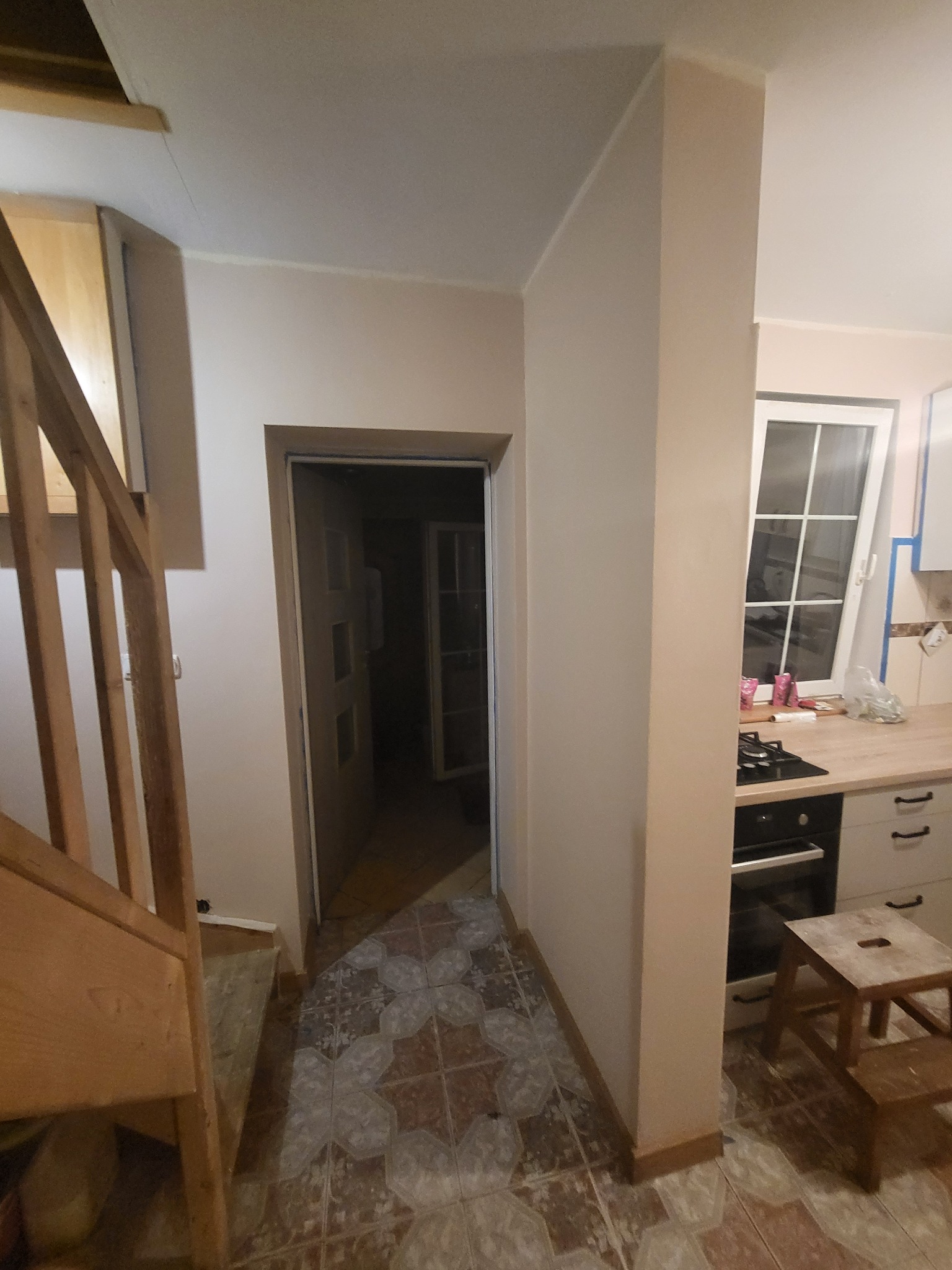 Wnętrze mieszkania w trakcie prac wykończeniowych: widok na korytarz z wzorzystą podłogą, otwarte drzwi do kolejnego pomieszczenia i fragment kuchni z oknem.