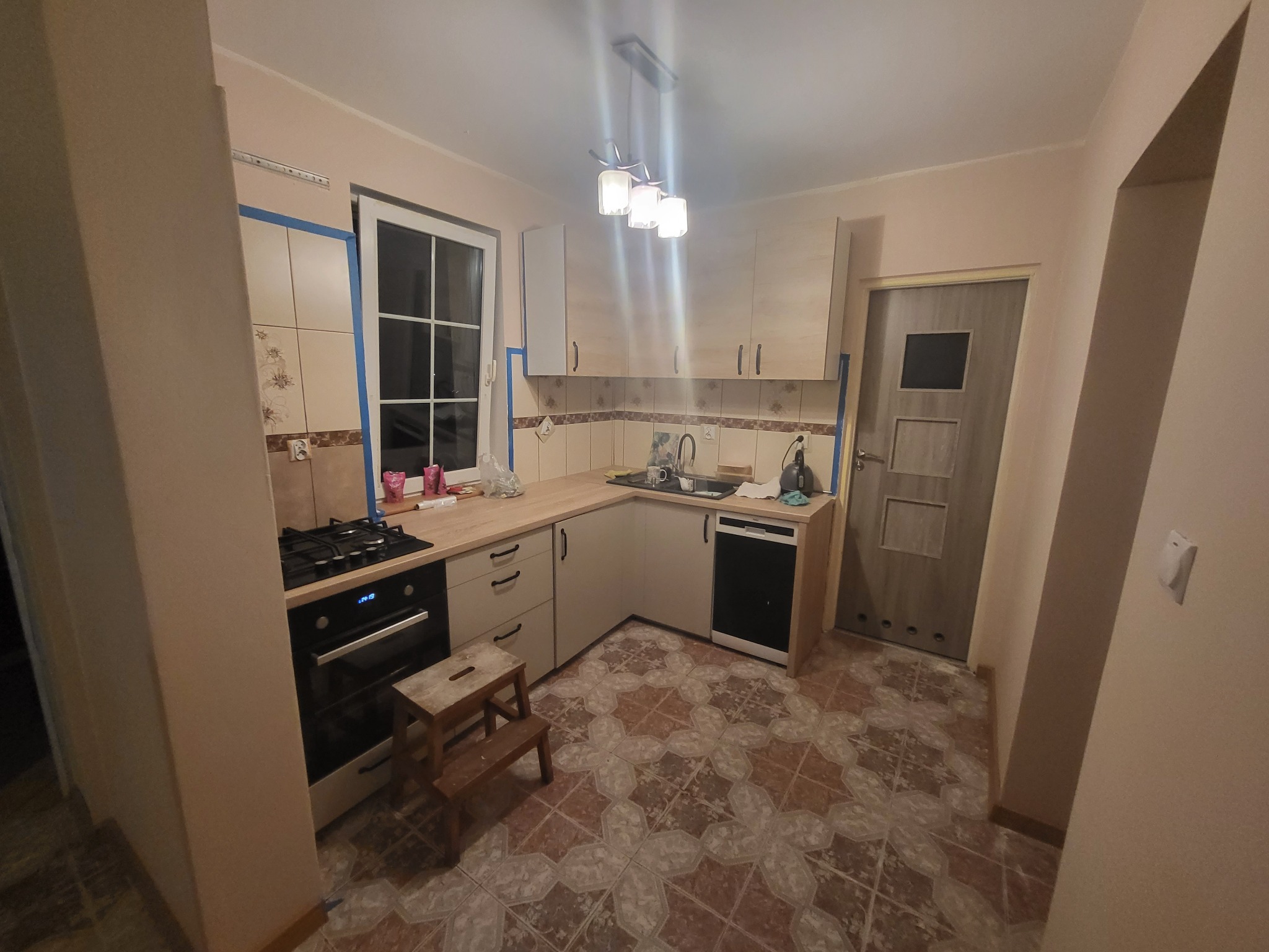 Kuchnia w trakcie prac wykończeniowych: widoczna zabudowa kuchenna z beżowymi frontami, kuchenka i piekarnik, płytki na ścianie zabezpieczone taśmą malarską, drabinka.