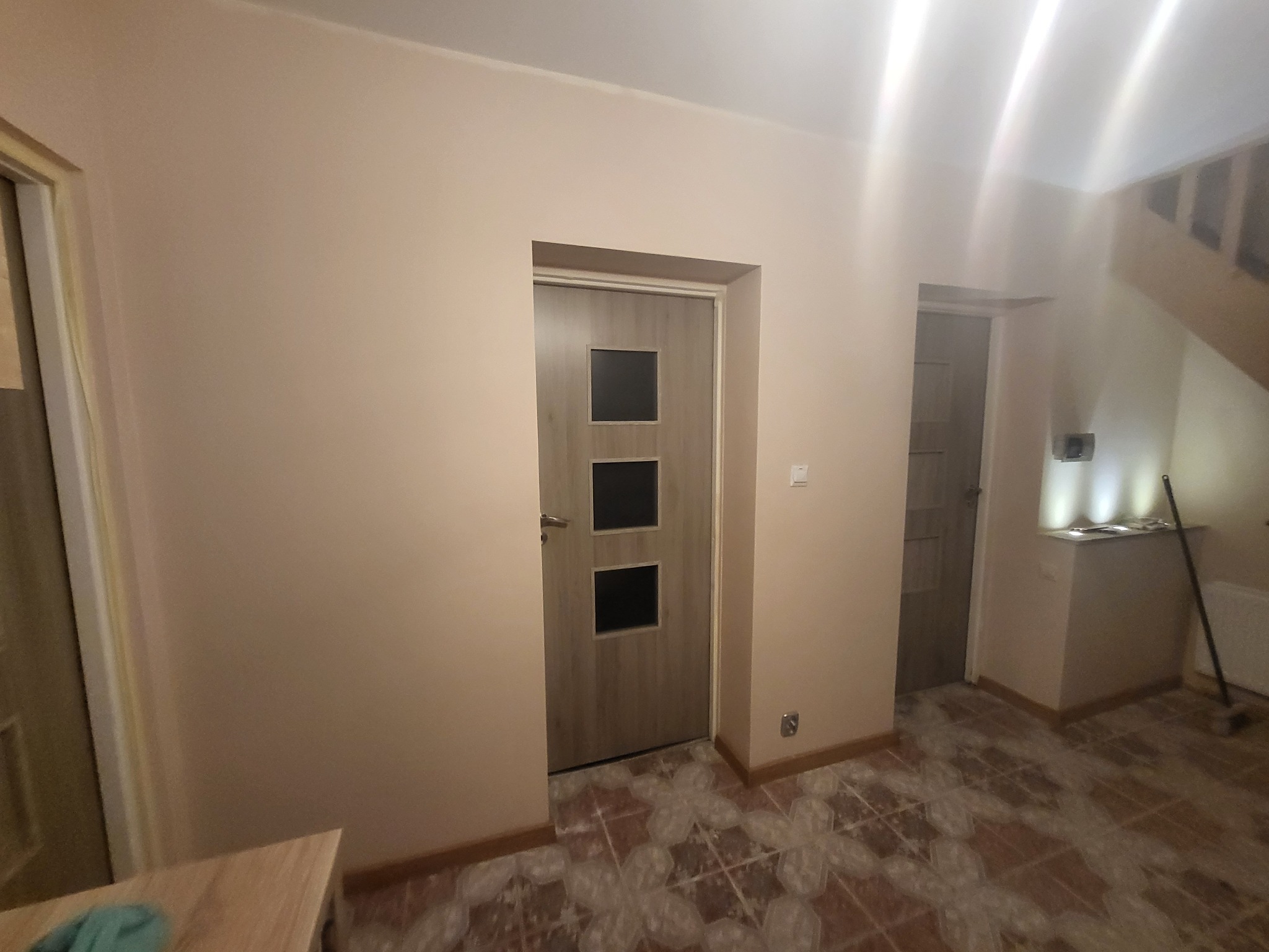 Wnętrze po odświeżeniu: beżowe ściany, szara, nowoczesna drzwi z trzema czarnymi szybkami, częściowo zakryta podłoga z wzorem w romby, widoczne schody w tle.