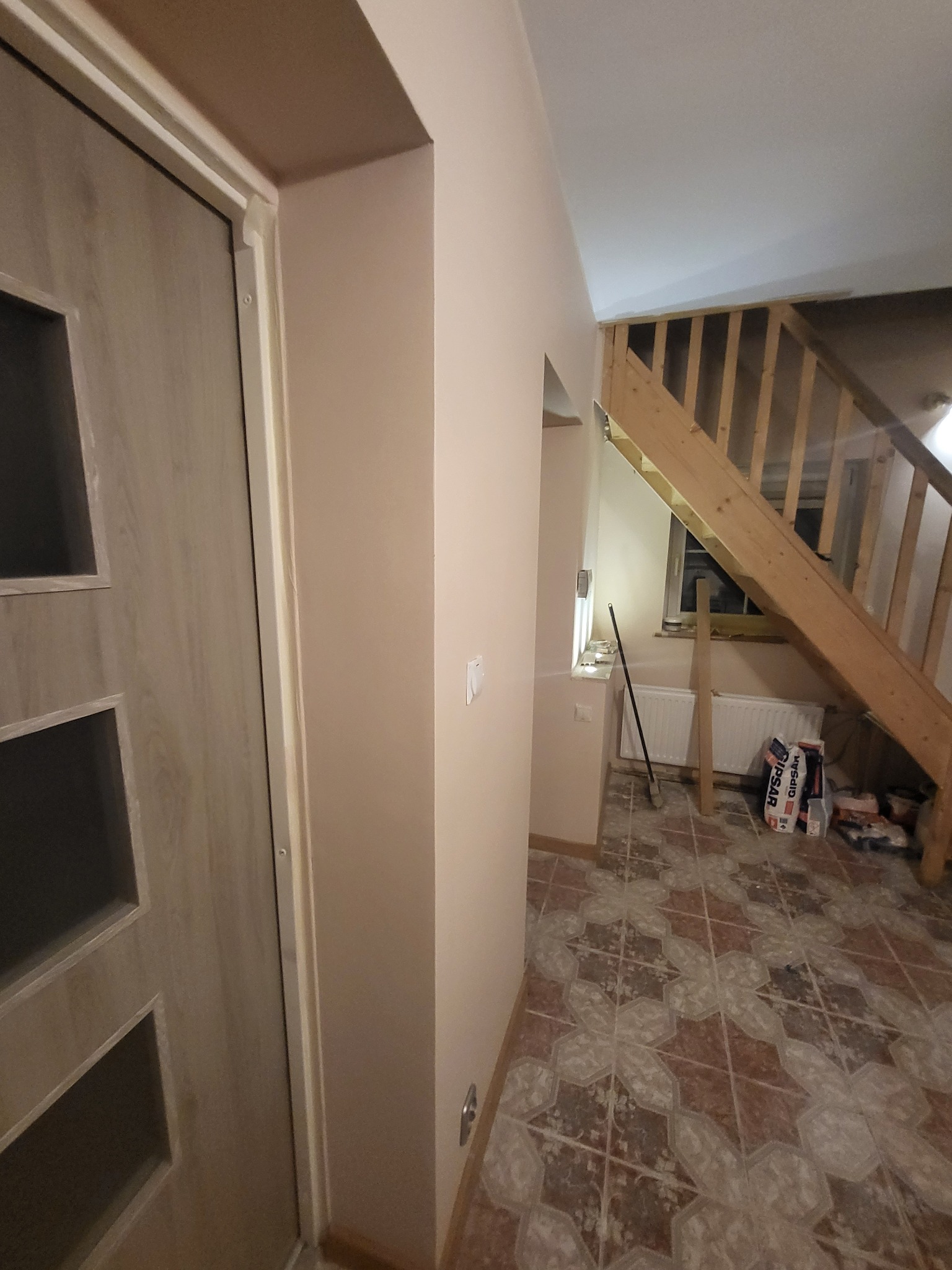 Wnętrze w trakcie prac wykończeniowych: widoczna drewniana klatka schodowa, drzwi wewnętrzne z przeszkleniami, fragment ściany pomalowanej na beżowo, podłoga z wzorzystej terakoty pokryta pyłem...