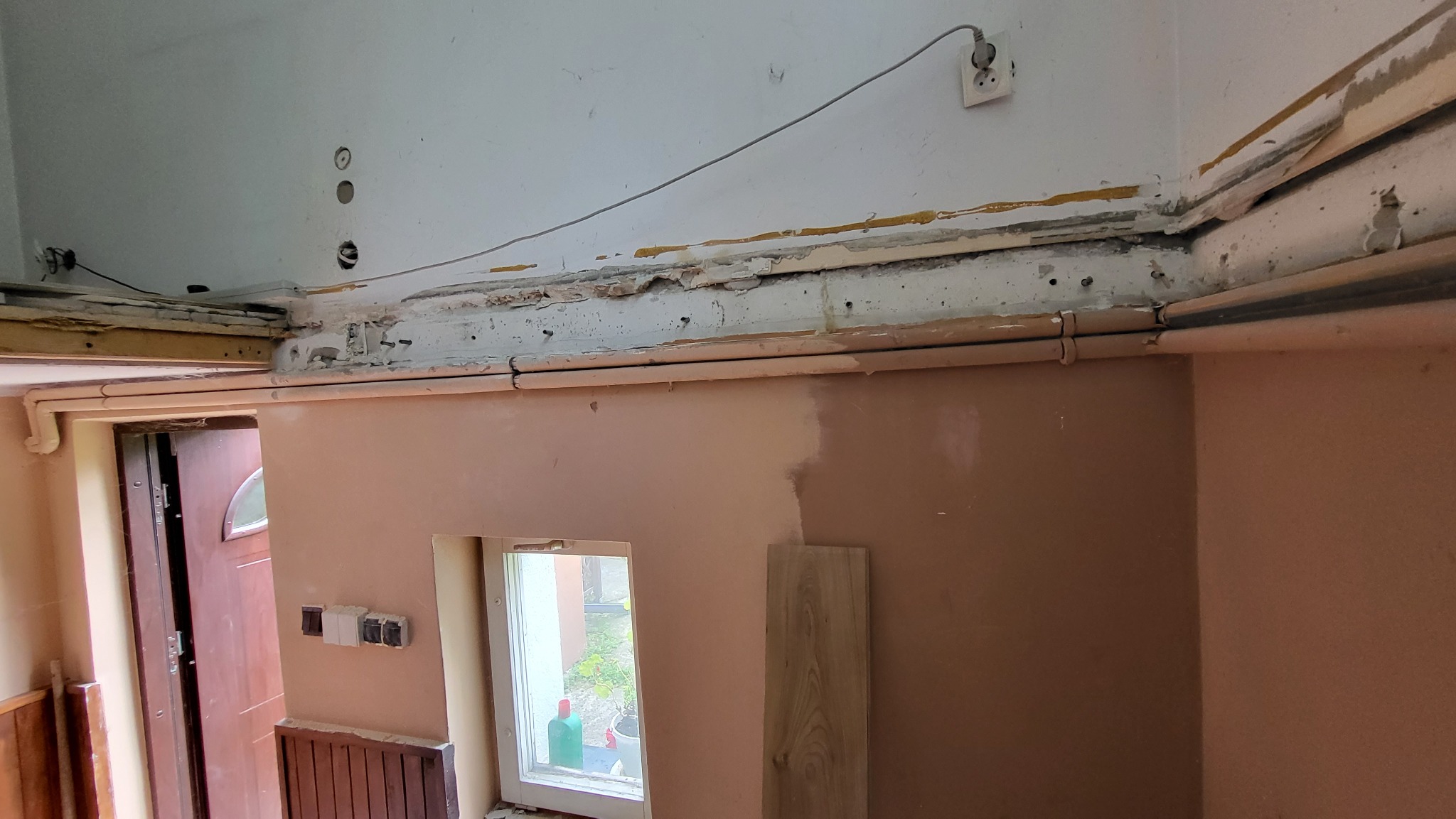 Wnętrze w trakcie remontu: ściany częściowo pokryte świeżym tynkiem, widoczne stare instalacje elektryczne, rury grzewcze i fragment okna z zielenią na zewnątrz.