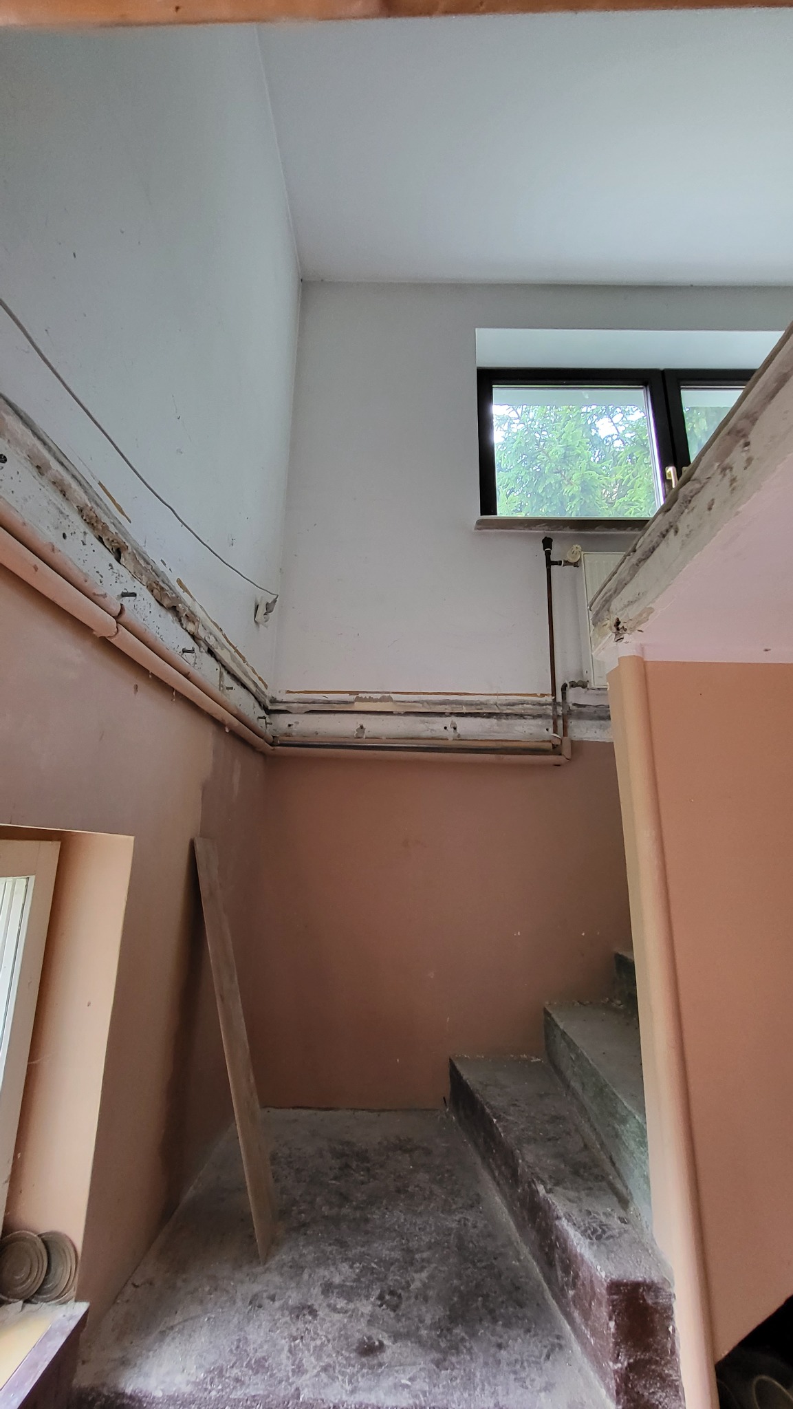 Klatka schodowa w trakcie prac przygotowawczych: odsłonięte rury grzewcze, usunięte listwy przypodłogowe, widoczne ślady po starych instalacjach, surowe ściany w odcieniach beżu i bieli, okno...
