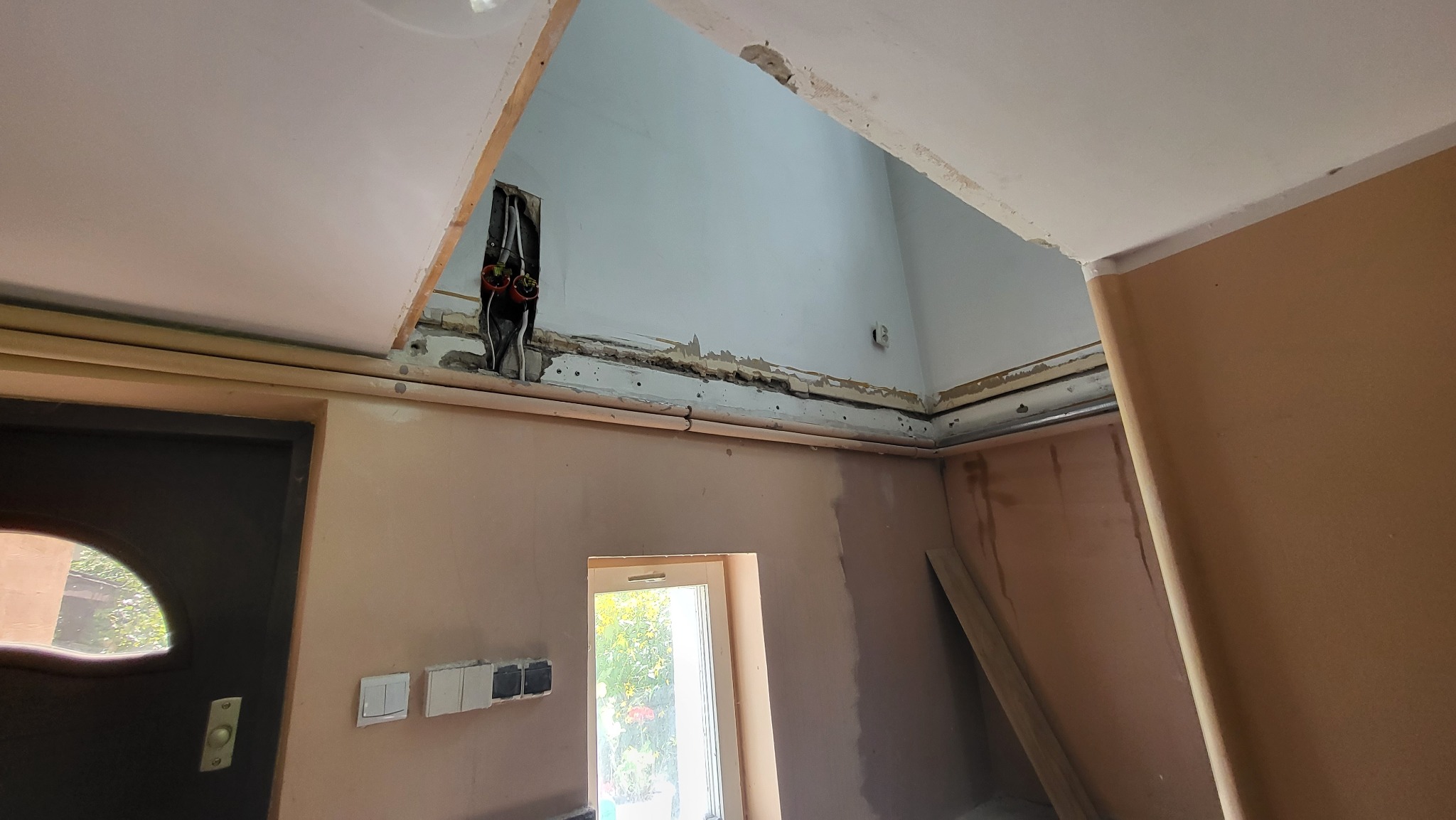 Wnętrze w trakcie prac przygotowawczych do wykończenia: odsłonięte przewody elektryczne w suficie, częściowo otynkowane ściany, widoczna instalacja pod sufitem i fragment okna.