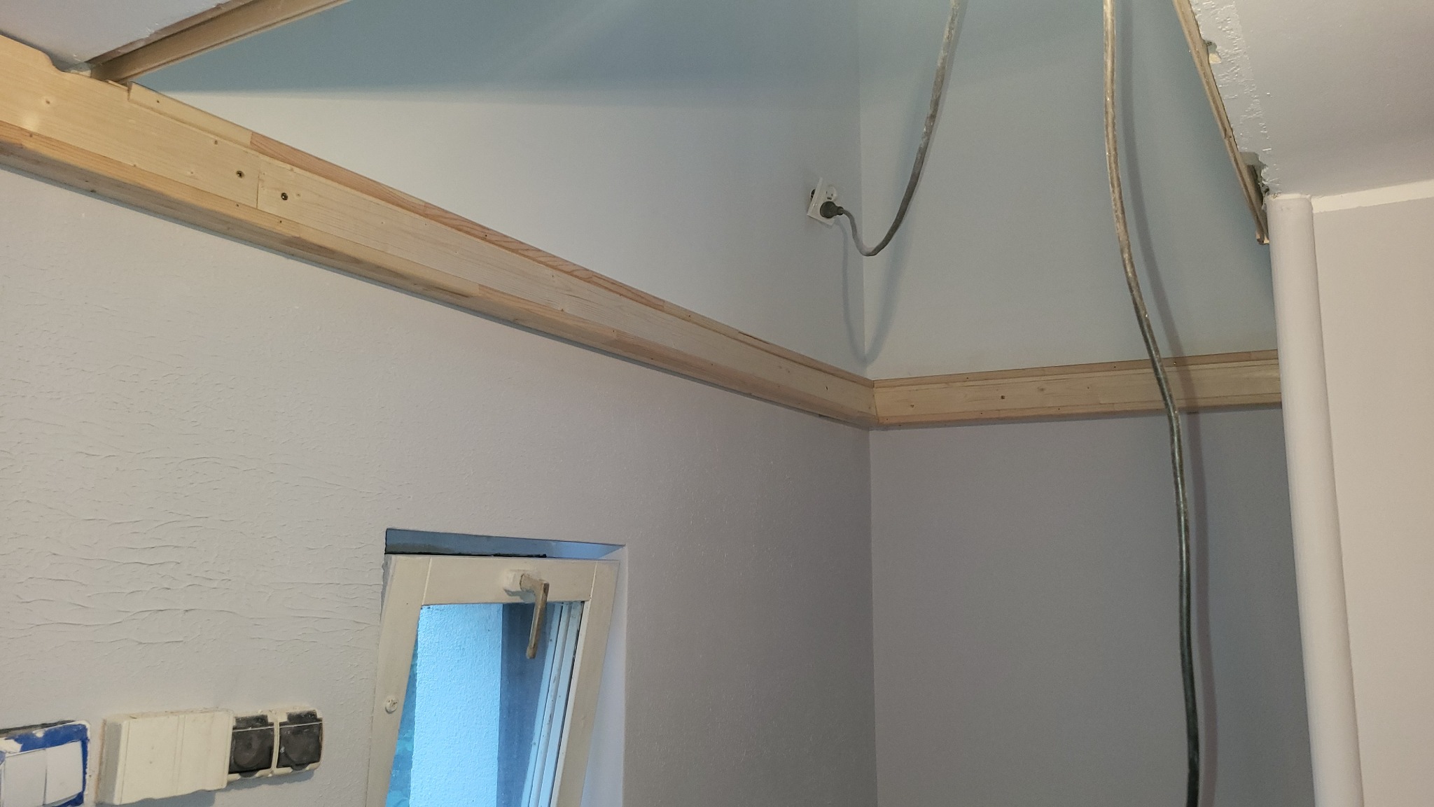 Surowe drewniane listwy zamontowane pod sufitem, widoczne kable wystające ze ściany i otwarte okno w pomieszczeniu w trakcie prac wykończeniowych.