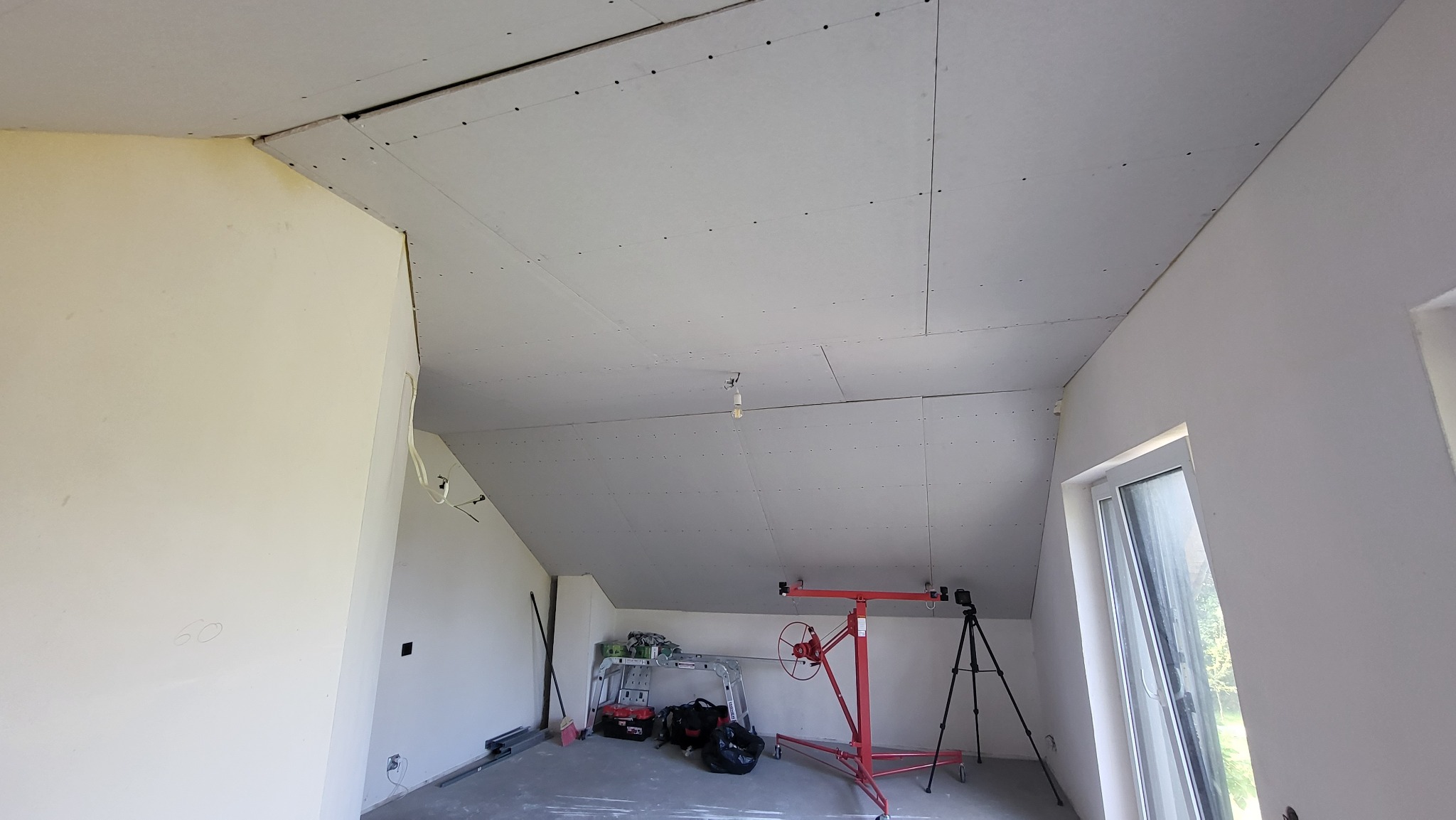 Płyty gipsowo-kartonowe zamontowane na suficie poddasza, widoczny podnośnik do płyt i inne narzędzia, surowy stan przed wykończeniem.