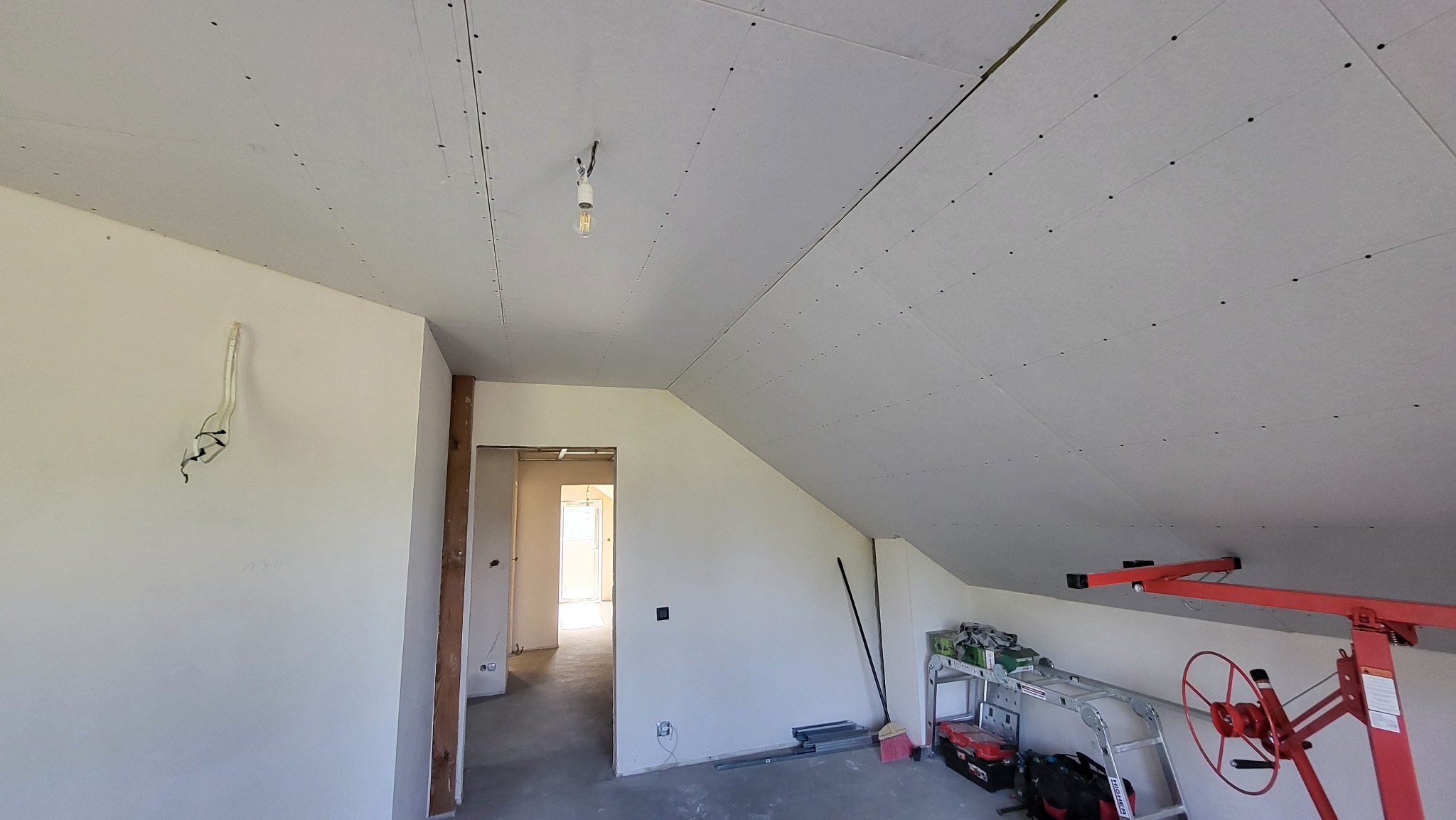 Poddasze w trakcie prac wykończeniowych: zamontowane płyty gipsowo-kartonowe na skosach i suficie, widoczny podnośnik do płyt, narzędzia na regale i wystające kable ze ściany.