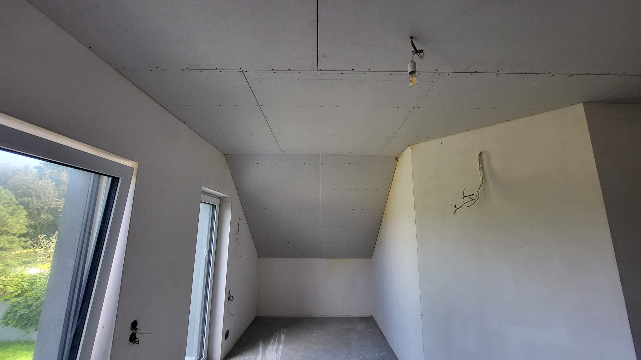 Pokój na poddaszu w trakcie przygotowywania do szpachlowania, z widocznymi płytami gipsowo-kartonowymi na suficie i ścianach, surowym betonem na podłodze, wiszącą żarówką na kablu oraz wystającymi...