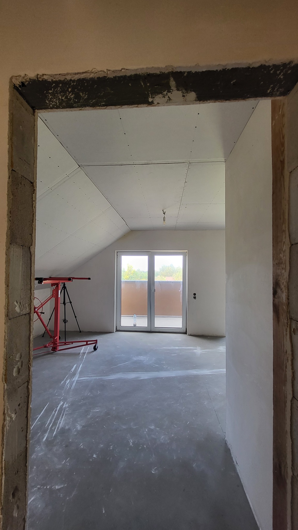 Wnętrze poddasza w trakcie prac wykończeniowych, widok przez surową futrynę, widoczny podnośnik do płyt gipsowych i wstępnie przygotowane ściany.