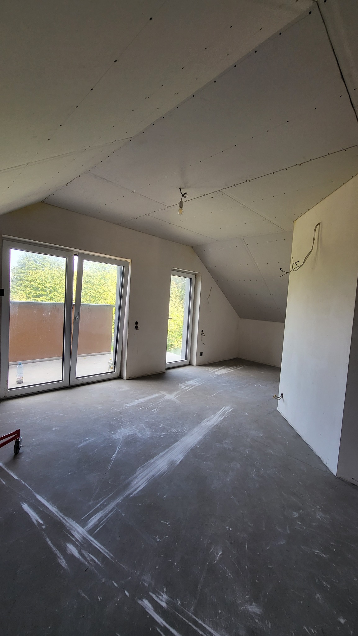 Poddasze w trakcie prac wykończeniowych: sufit z płyt gipsowo-kartonowych, szpachlowana podłoga, okna balkonowe z widokiem na zieleń.