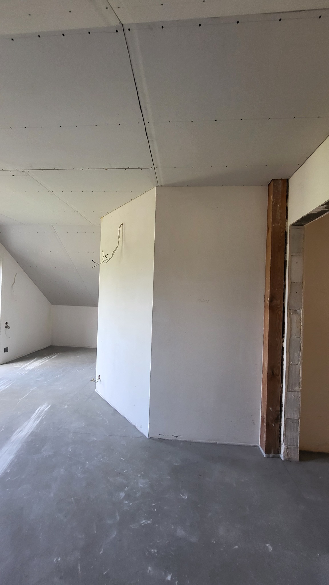 Surowe wnętrze poddasza z wykończonymi płytami gipsowo-kartonowymi na skosach i ścianach, widoczne kable elektryczne wystające ze ściany, surowa podłoga i fragment ościeżnicy drzwiowej.