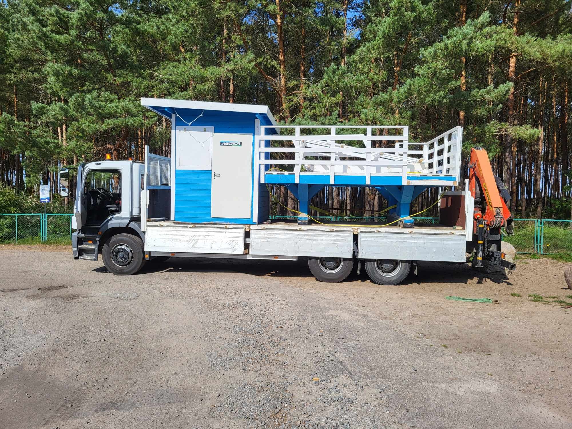 Ciężarówka z platformą załadunkową i dźwigiem przewozi niebiesko-białą konstrukcję domku z balustradą na tle lasu.