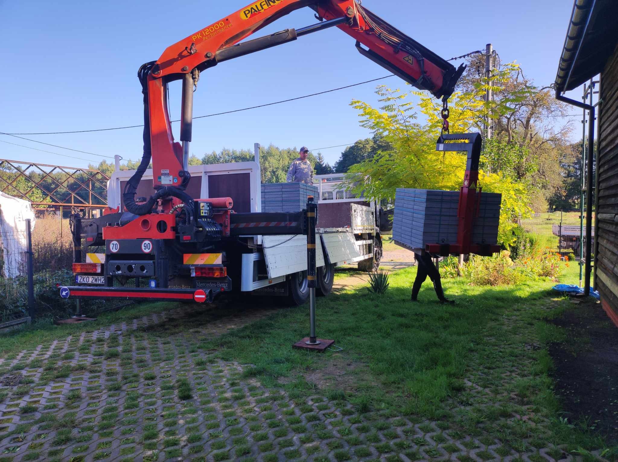 Czerwony dźwig Palfinger PK 12000 Performance zamontowany na ciężarówce Mercedes-Benz, podnosi paletę z szarymi płytami chodnikowymi na tle zieleni i błękitnego nieba.