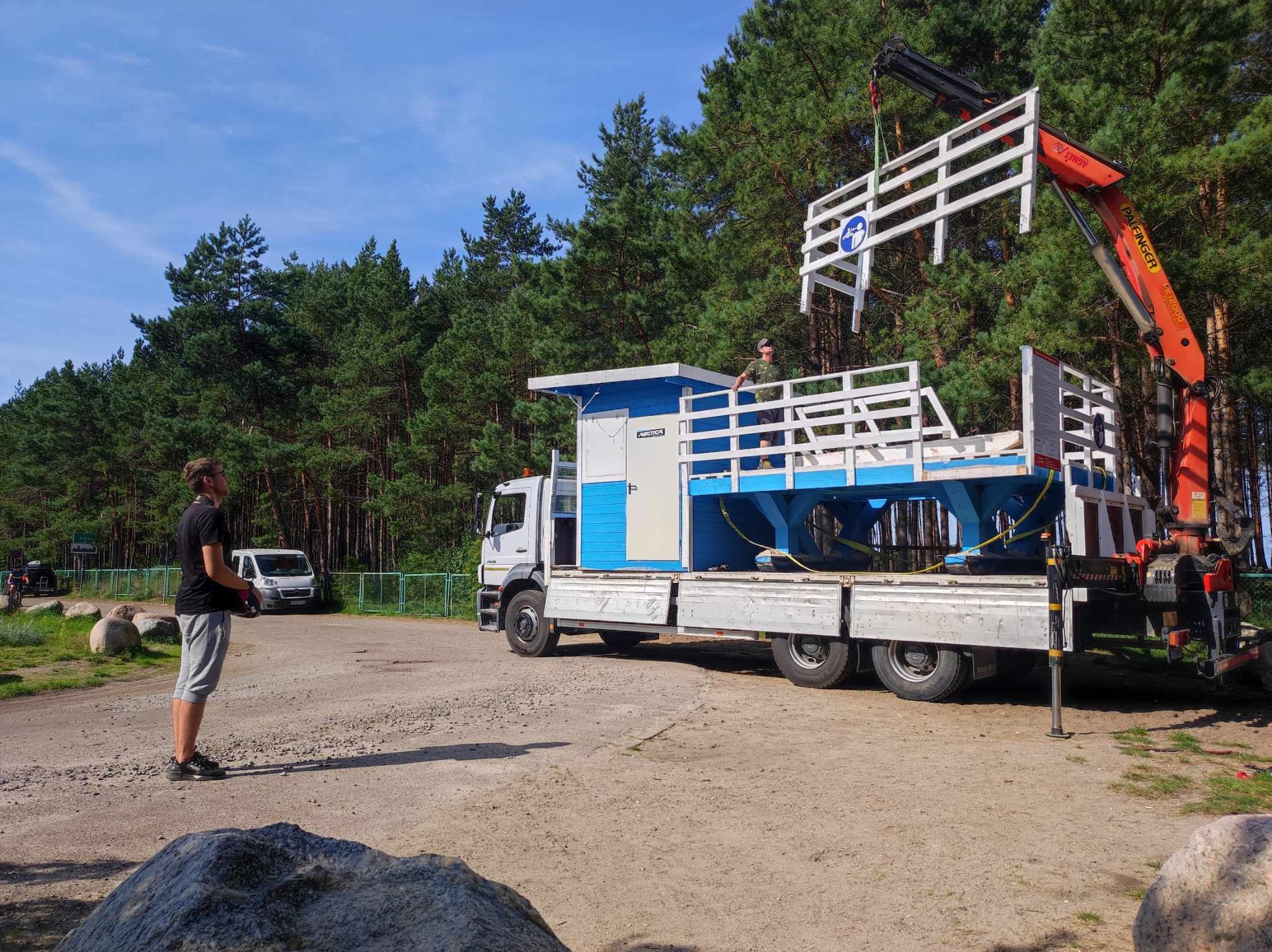 Ciężarówka z dźwigiem Palfinger podnosi element konstrukcyjny z białymi barierkami, prawdopodobnie fragment pomostu lub platformy widokowej, na tle lasu i błękitnego nieba. Operator na platformie...