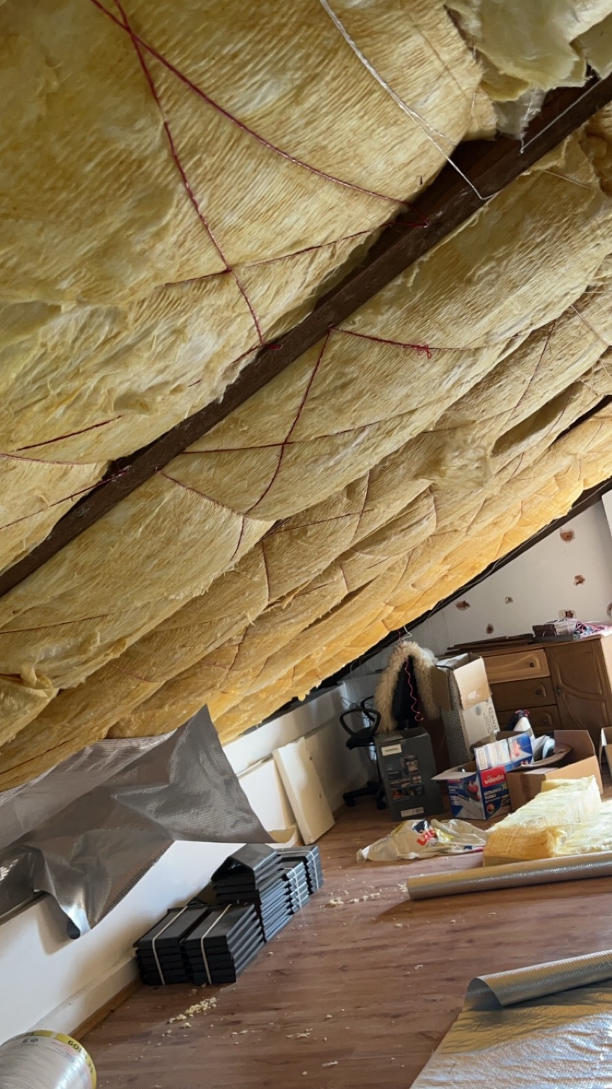 Izolacja poddasza wełną mineralną, zabezpieczona sznurkiem, widok z dołu, w tle fragment pokoju z meblami i kartonami.