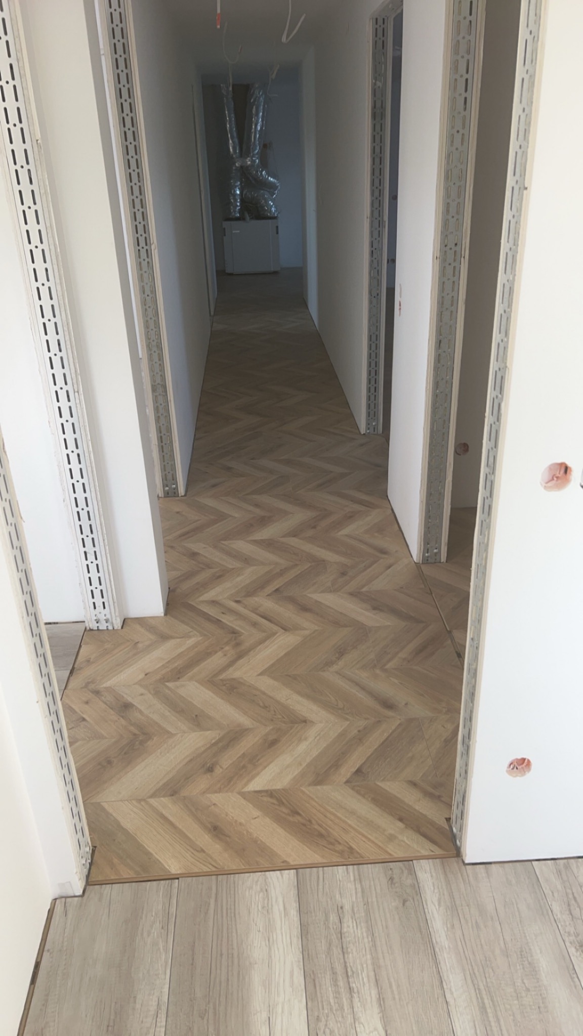 Ułożona parkietowa podłoga w jodełkę w korytarzu w trakcie remontu, widoczne metalowe profile przy ścianach i instalacja wentylacyjna w tle.