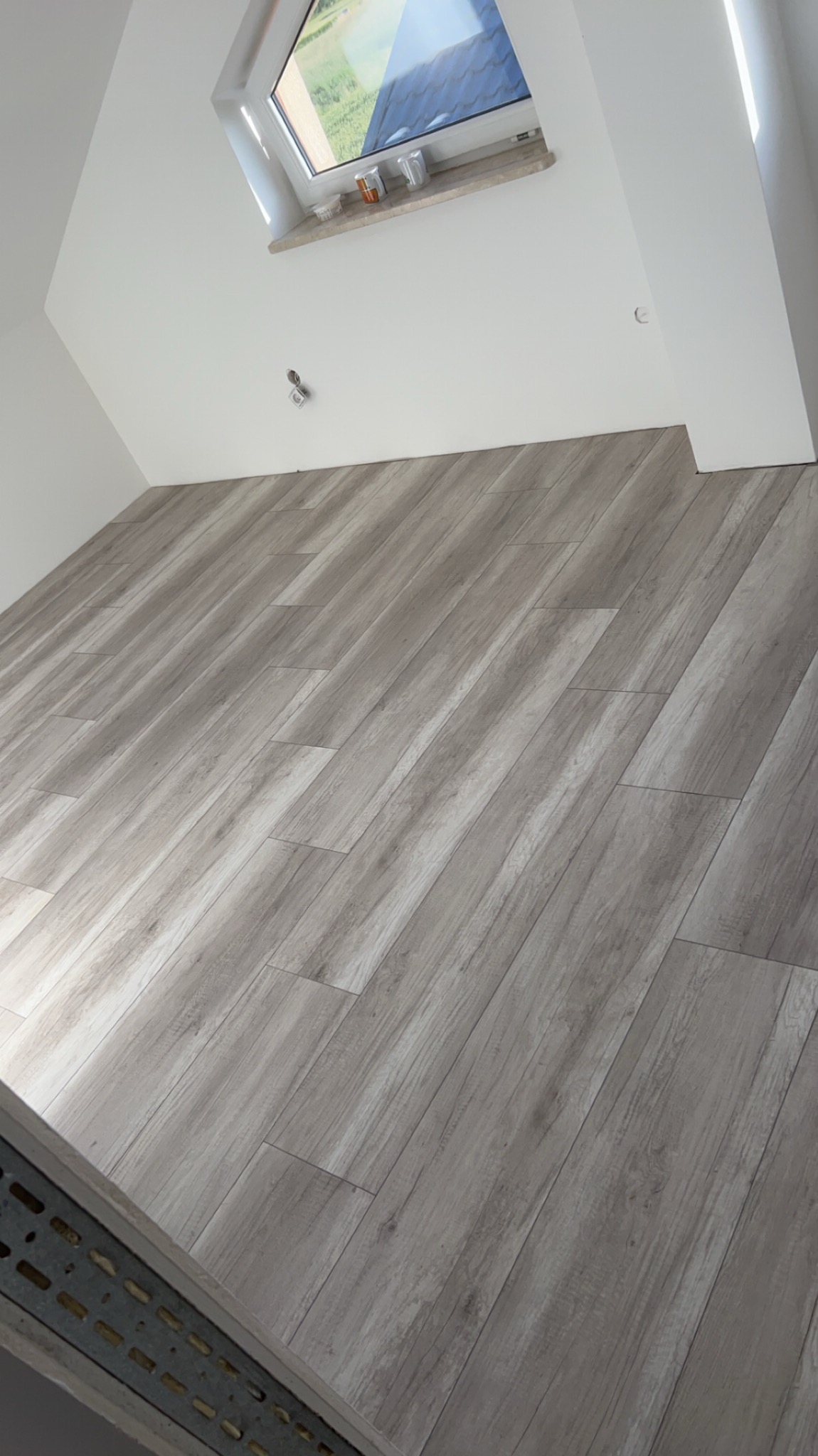 Ukończona podłoga z paneli laminowanych w odcieniach szarości, ułożona w skos w jasnym pokoju na poddaszu z oknem dachowym.